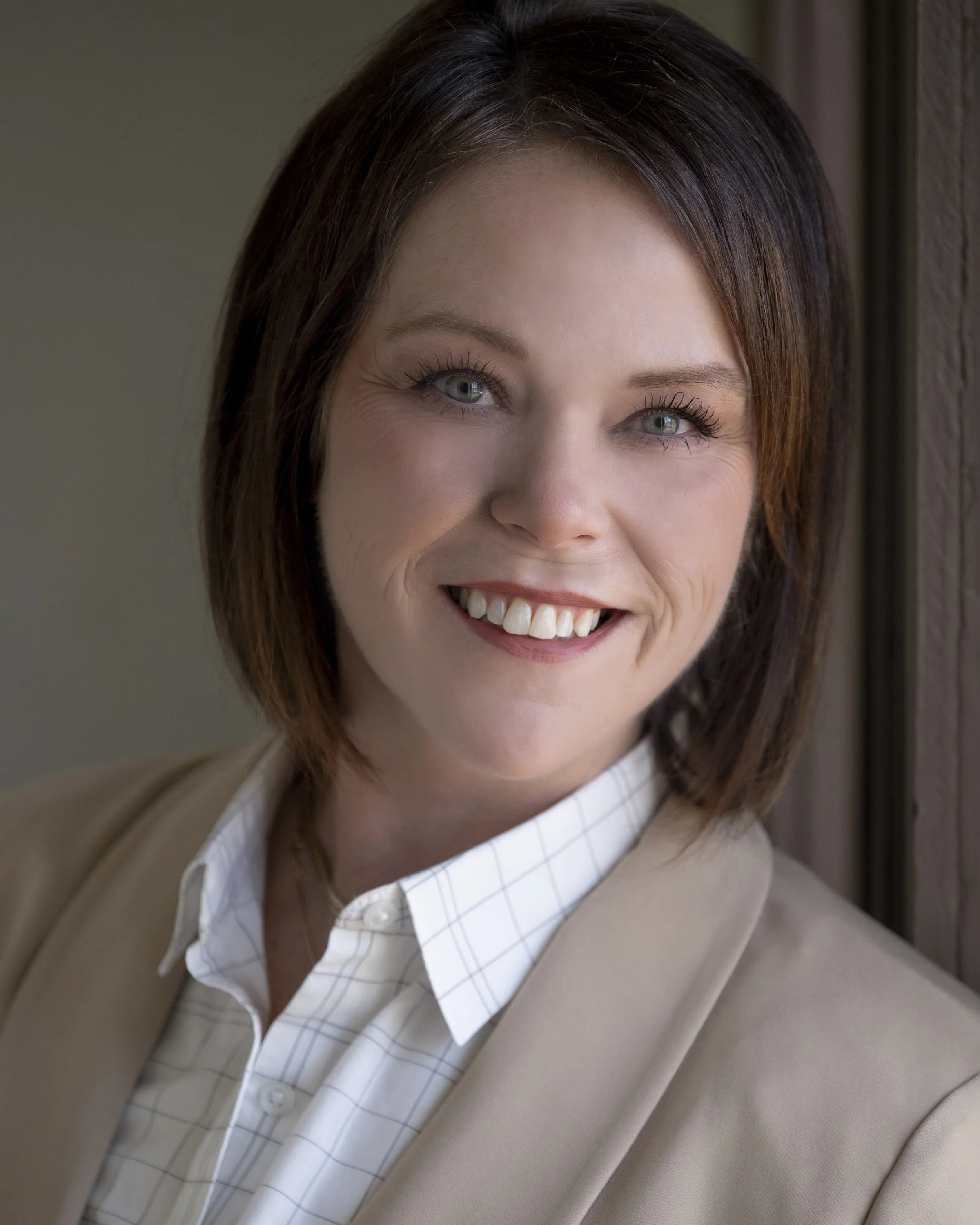 A woman with short brown hair, blue eyes, and a bright smile, wearing a beige blazer and a white checked shirt, standing near a window.