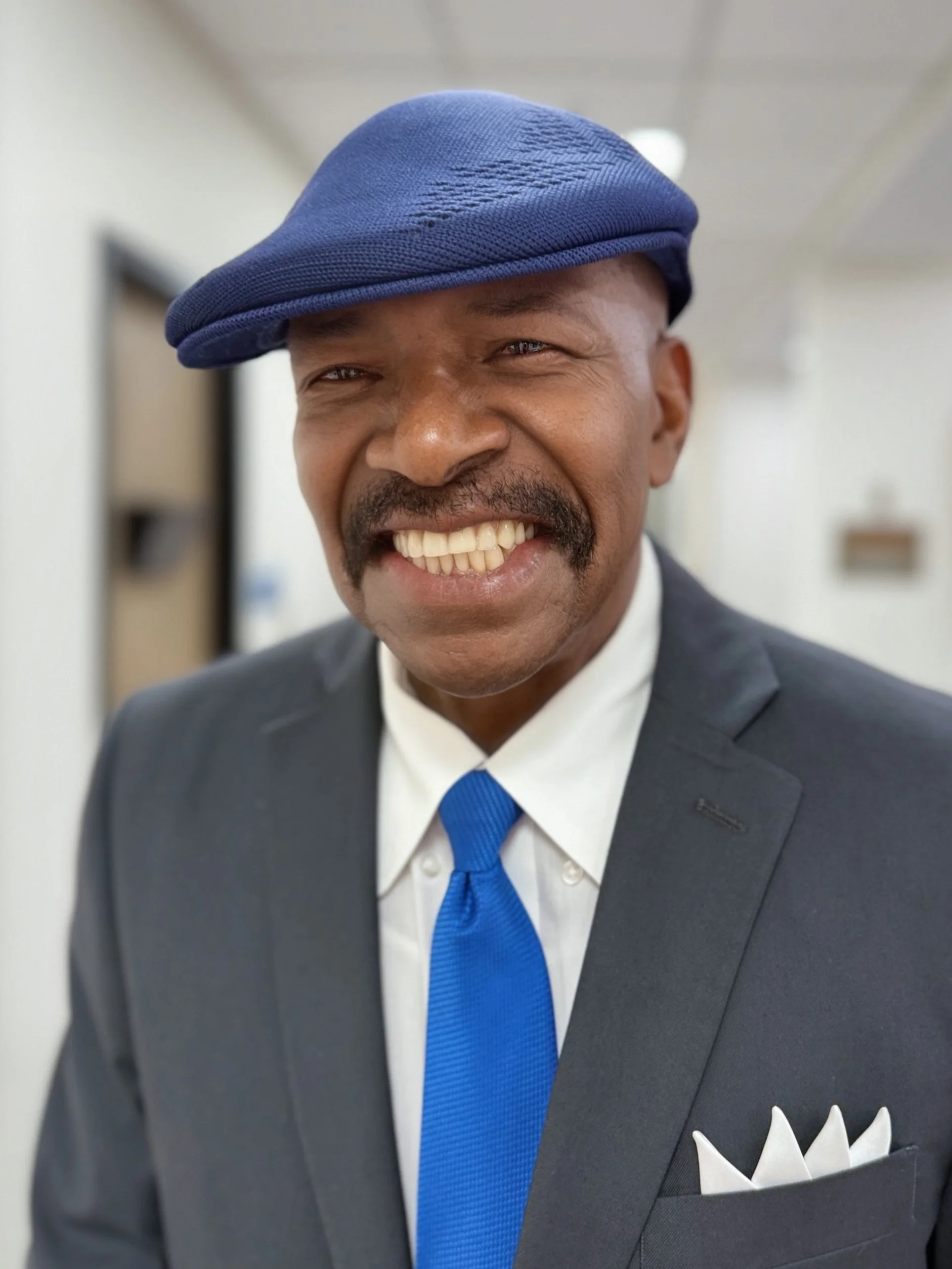 A smiling man wearing a dark suit, white shirt, blue tie, and a blue beret.