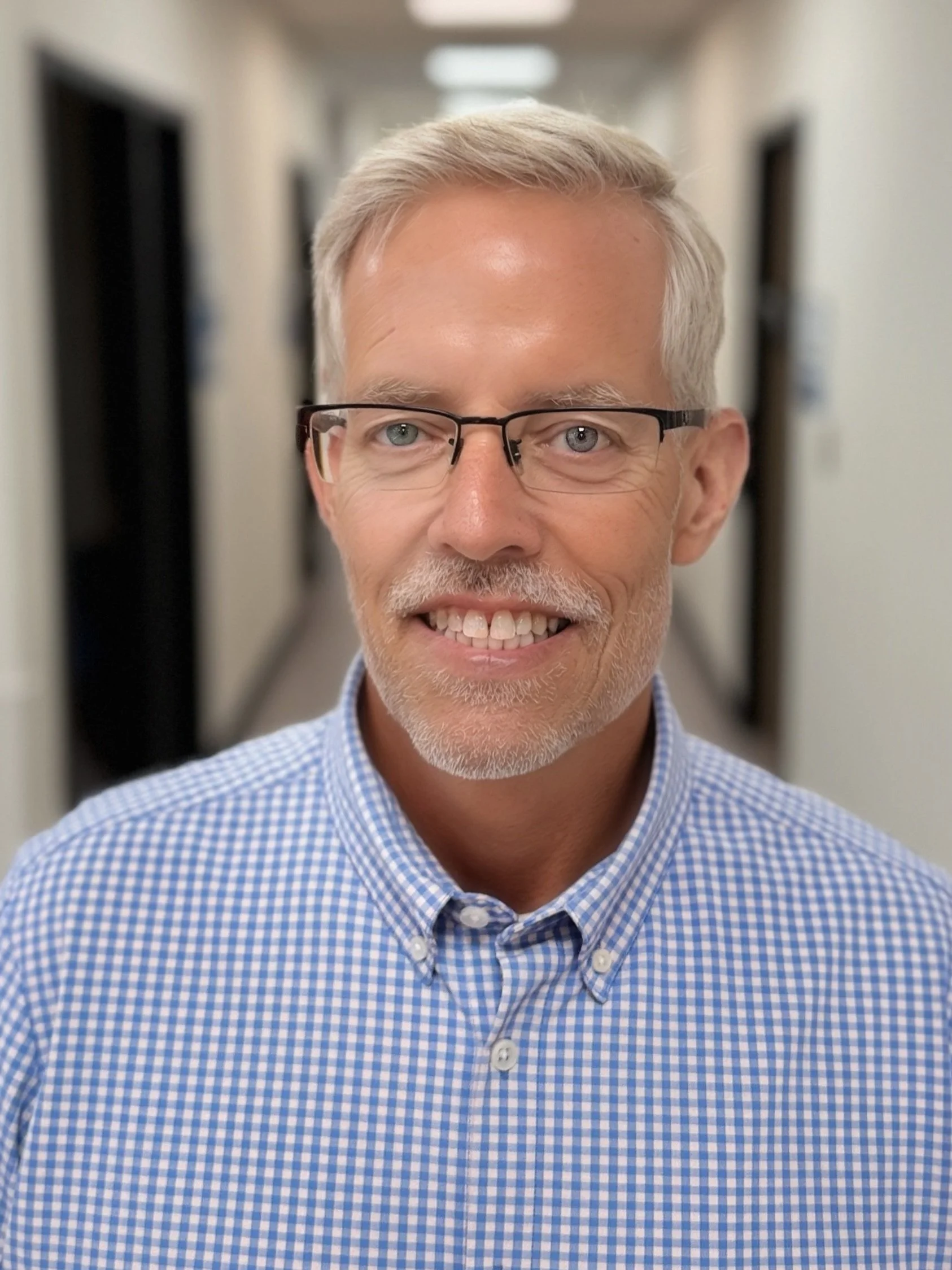 A middle-aged man with gray hair, glasses, and a beard, smiling while standing in a corridor wearing a blue checkered shirt.