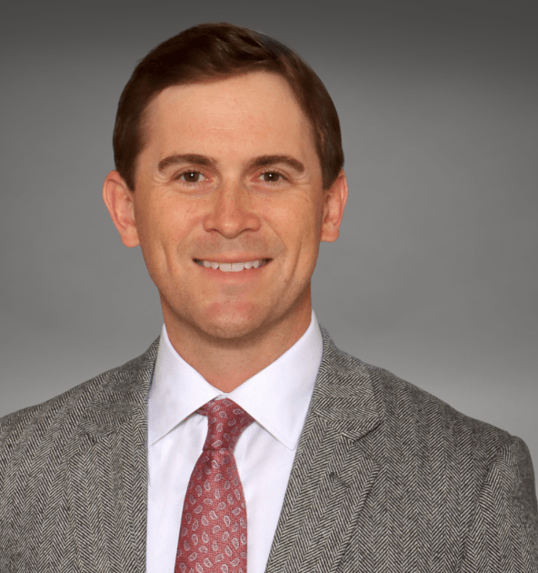 Portrait of a man in a gray herringbone suit, white shirt, and red patterned tie, smiling against a neutral gray background.