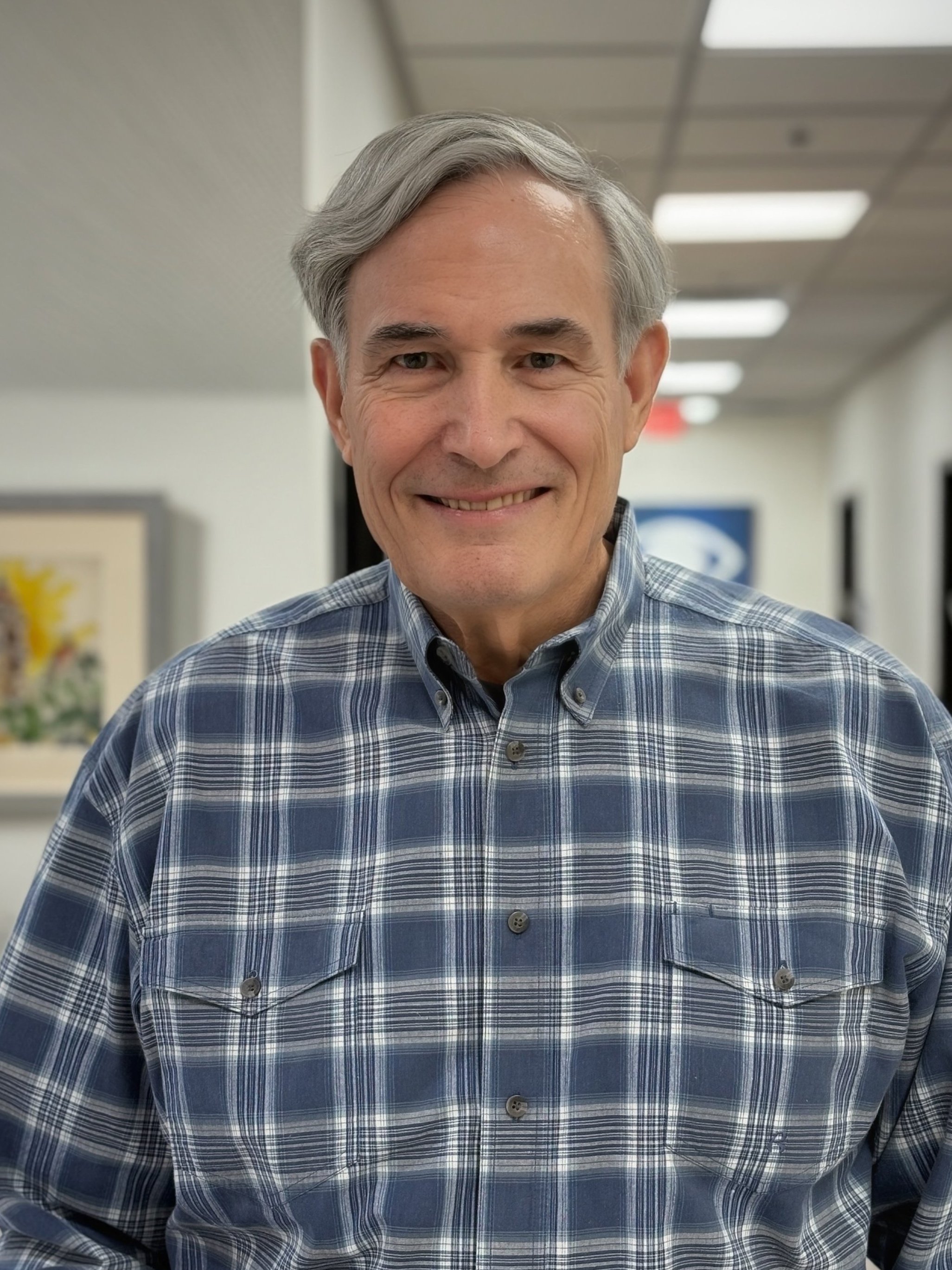 An older man with gray hair smiling, wearing a dark blazer over a checkered shirt, standing in an indoor setting with blurred people and doors in the background.