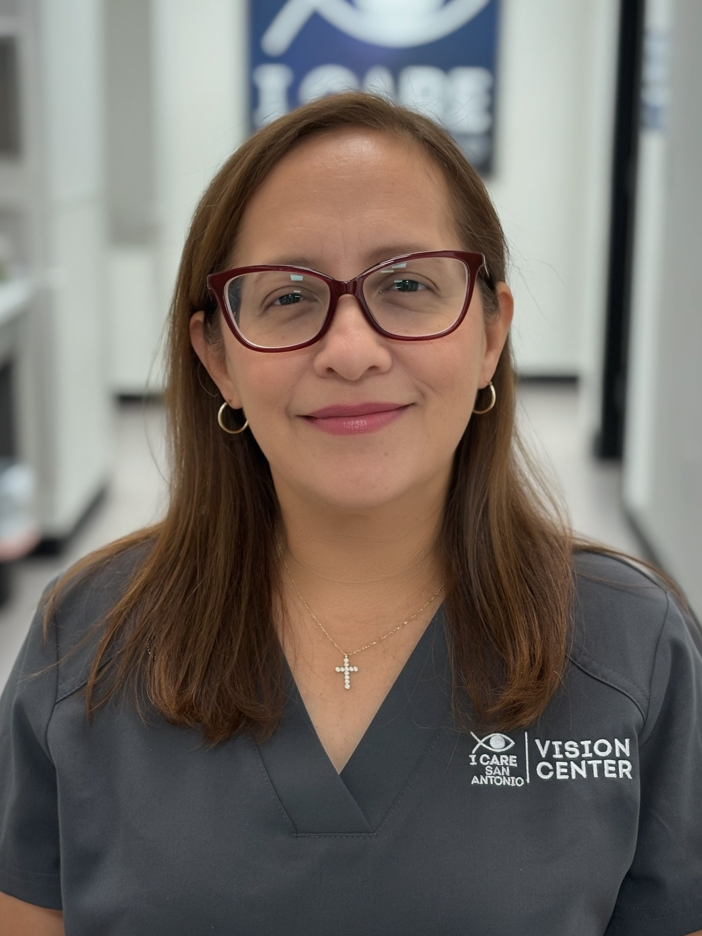 A woman with brown hair, wearing glasses, earrings, and a cross necklace, smiling at the camera in a medical or eye care center. She is dressed in a gray uniform with the logo for I Care San Antonio Vision Center.