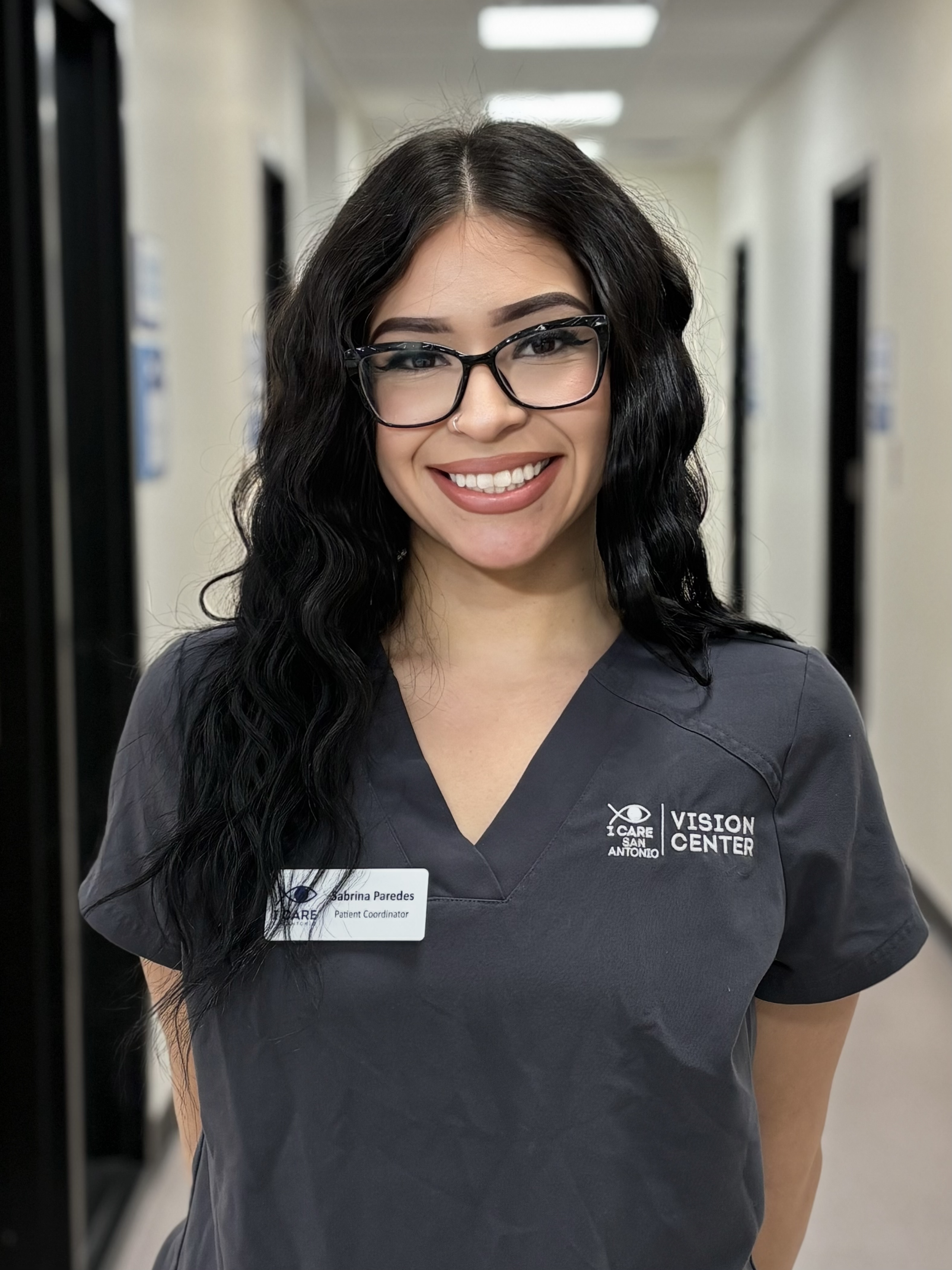 Smiling woman in grey scrubs with 'Vision Center' and 'I Care San Antonio' logos, standing in a clinical setting, wearing a cross necklace and colorful bracelets.