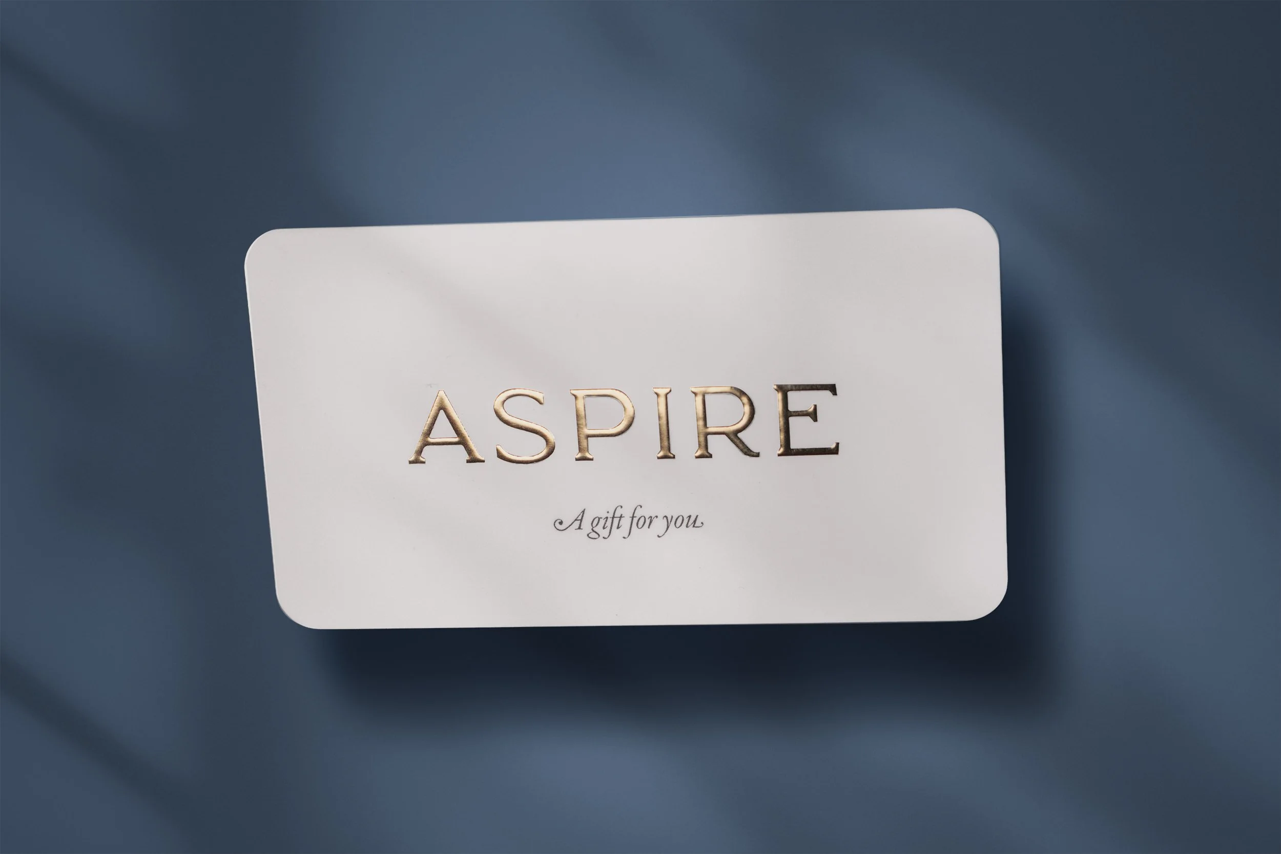 A white card with the word 'ASPIRE' in gold letters and the phrase 'A gift for you' written in cursive below, placed on a blue fabric background.