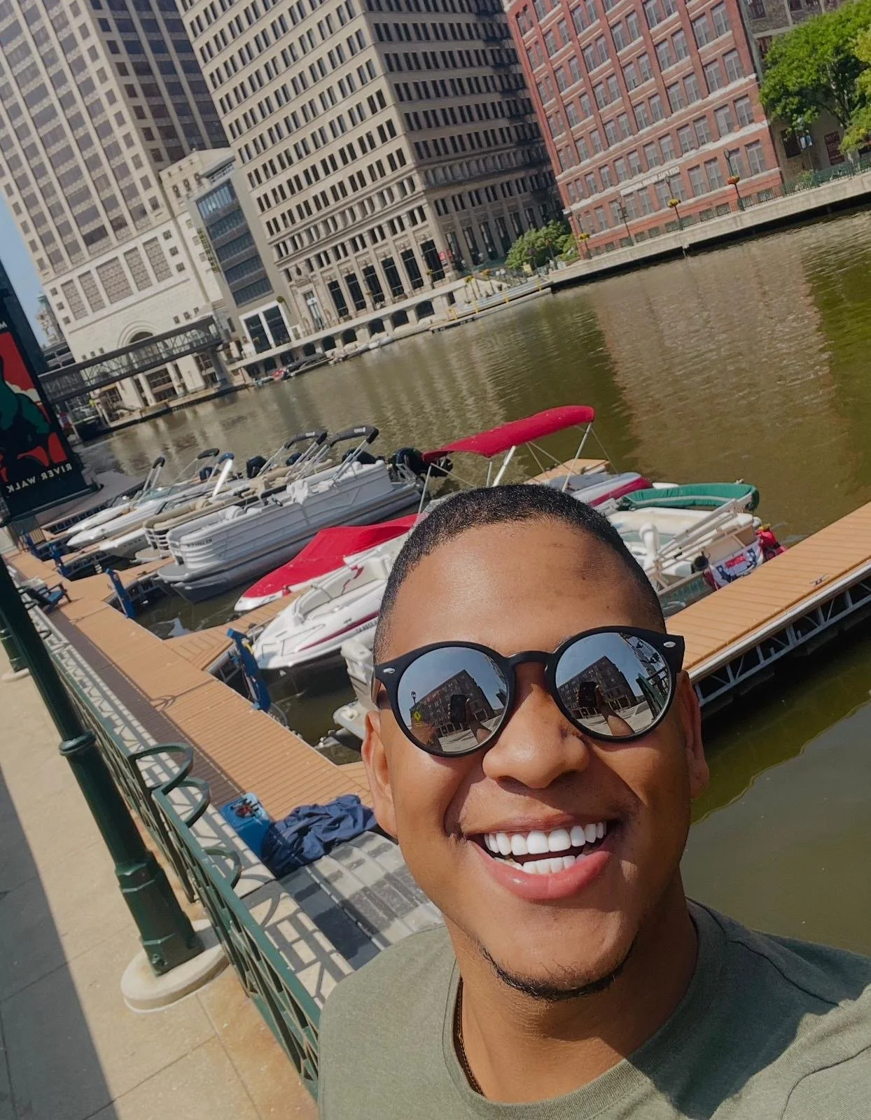 A smiling man with black sunglasses taking a selfie near a marina with boats, reflected buildings, and a cityscape in the background on a sunny day.