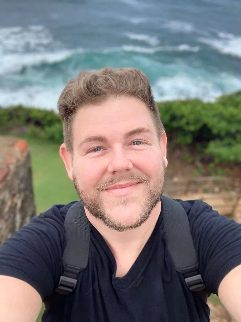 A man taking a selfie outdoors with ocean waves in the background, wearing a black shirt and carrying a backpack.