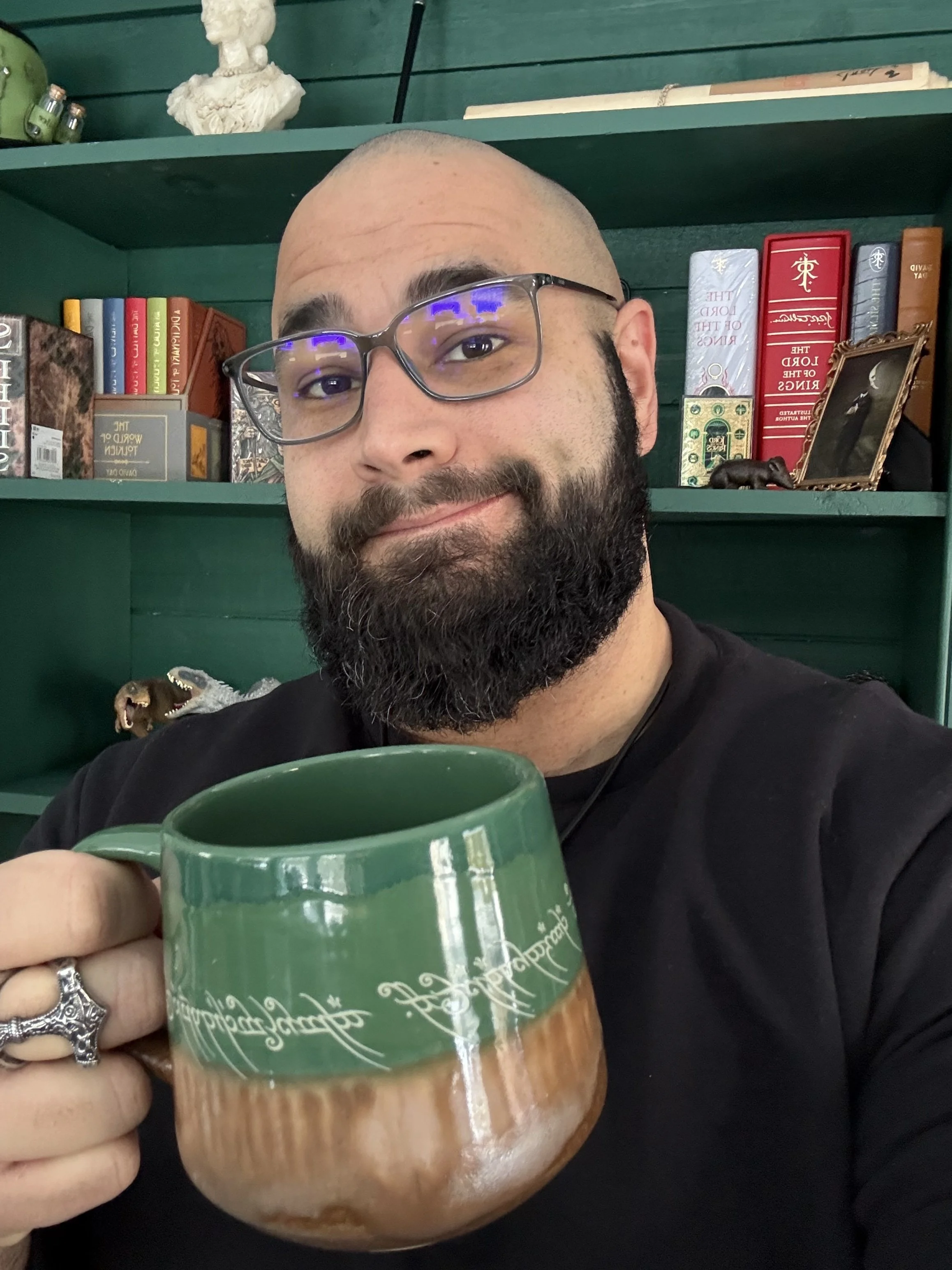 A man with glasses and a beard holding a green and brown ceramic mug, smiling in front of a bookshelf filled with books and decorative items.