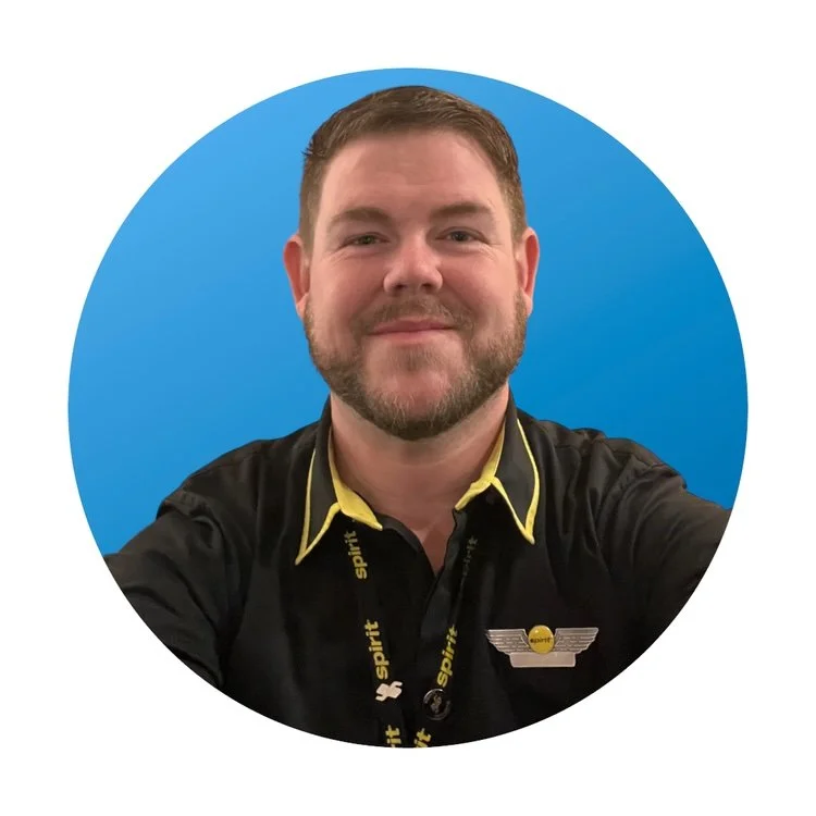 A man with a beard and short brown hair taking a selfie, wearing a black shirt with yellow accents and a spirit airline badge, standing in front of a blue circular background.