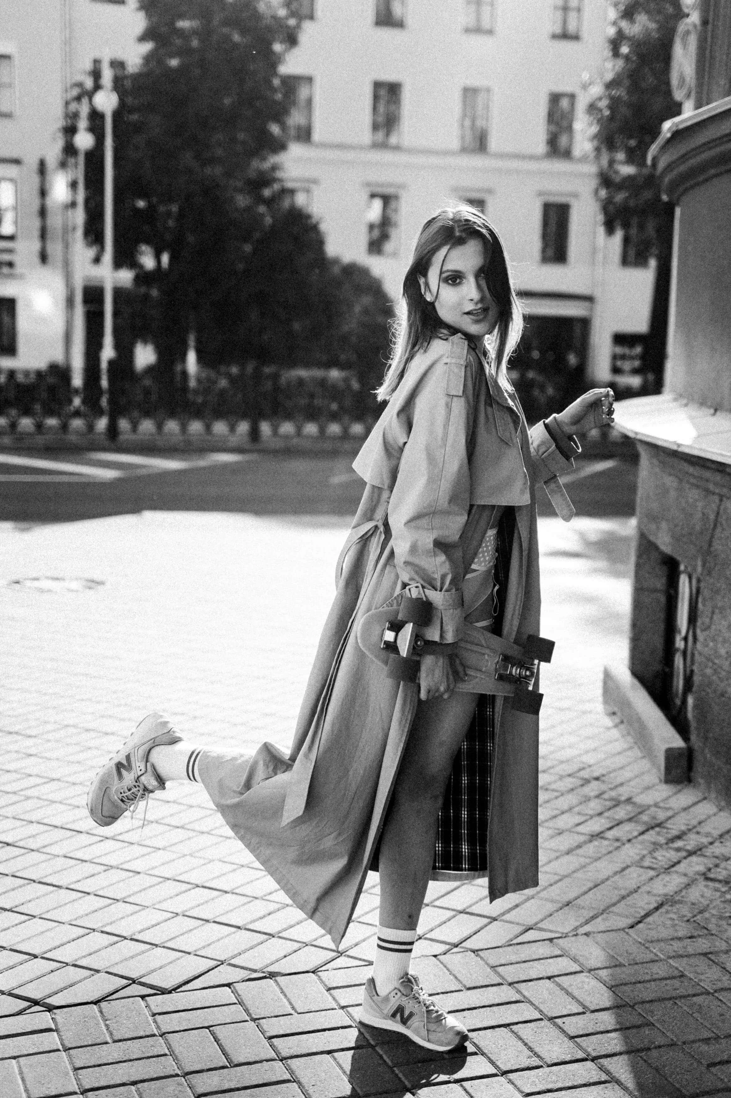 A young woman dressed in a trench coat and sneakers standing on a city sidewalk at dusk, holding a skateboard and looking at the camera.