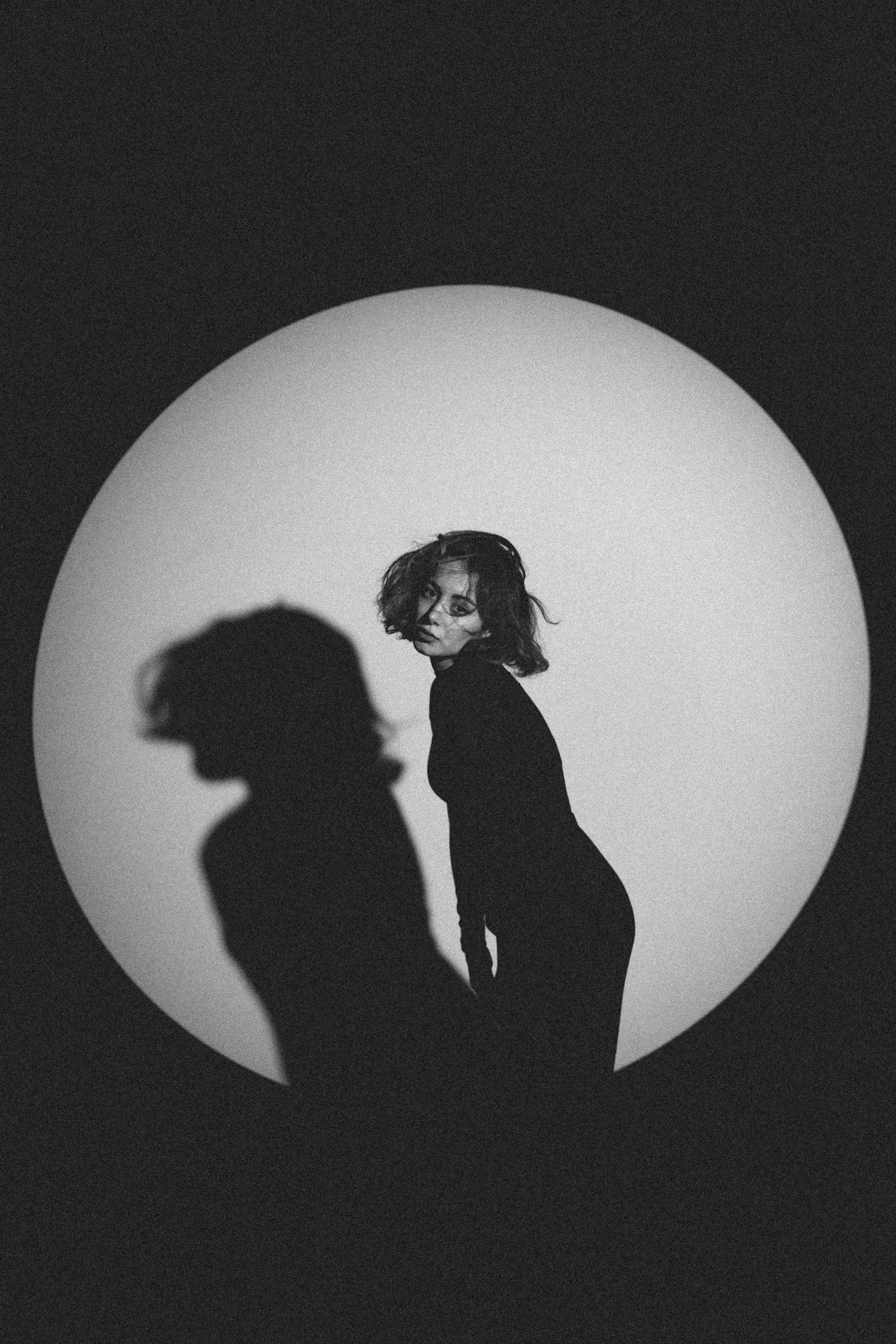 Black and white image of a woman with wavy hair standing in front of a circular light source. Her shadow is cast on the background, creating a silhouette. The woman is in a black outfit, looking over her shoulder.