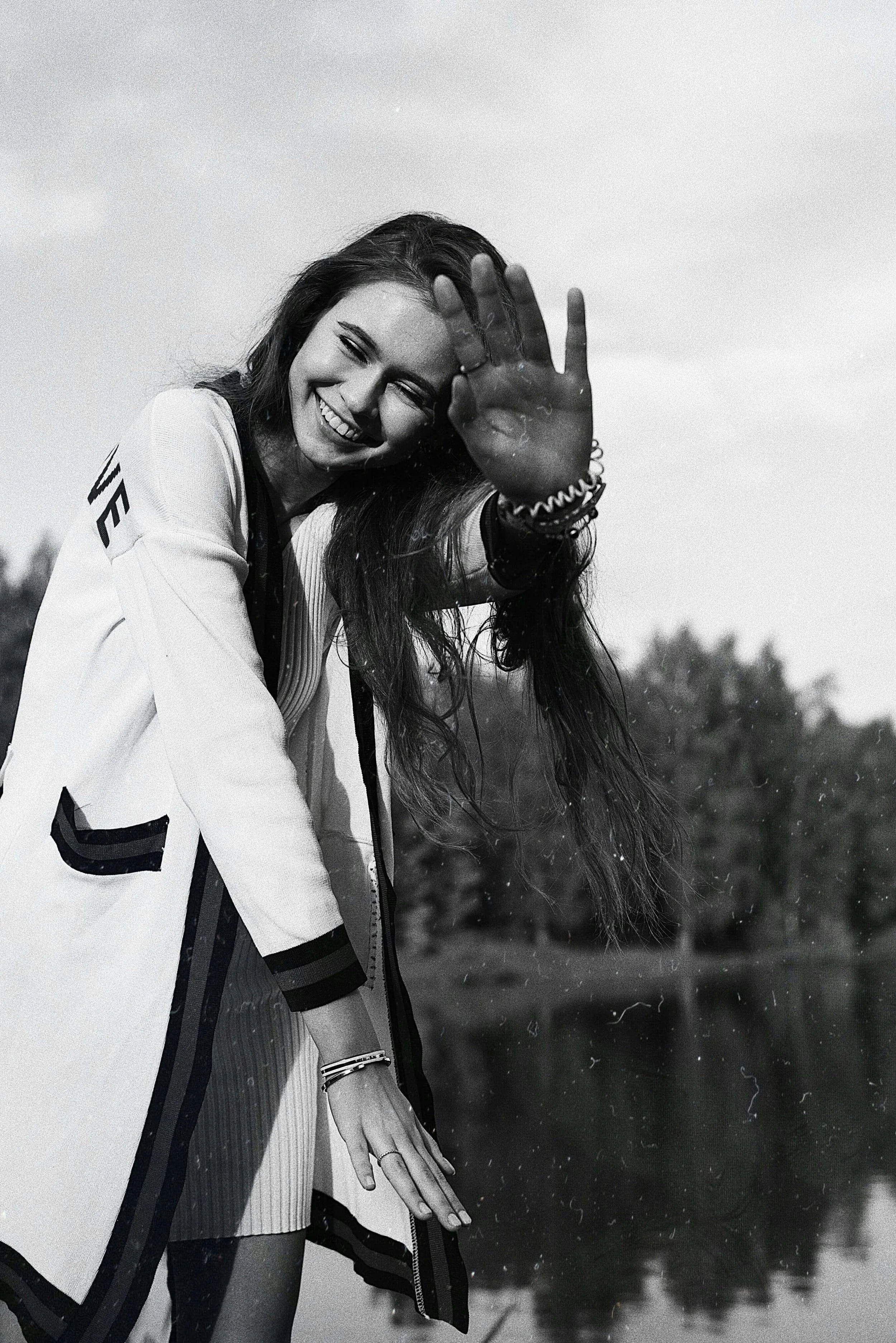 A smiling young woman with long hair reaching out towards the camera, her hand in focus, outdoors near a body of water.