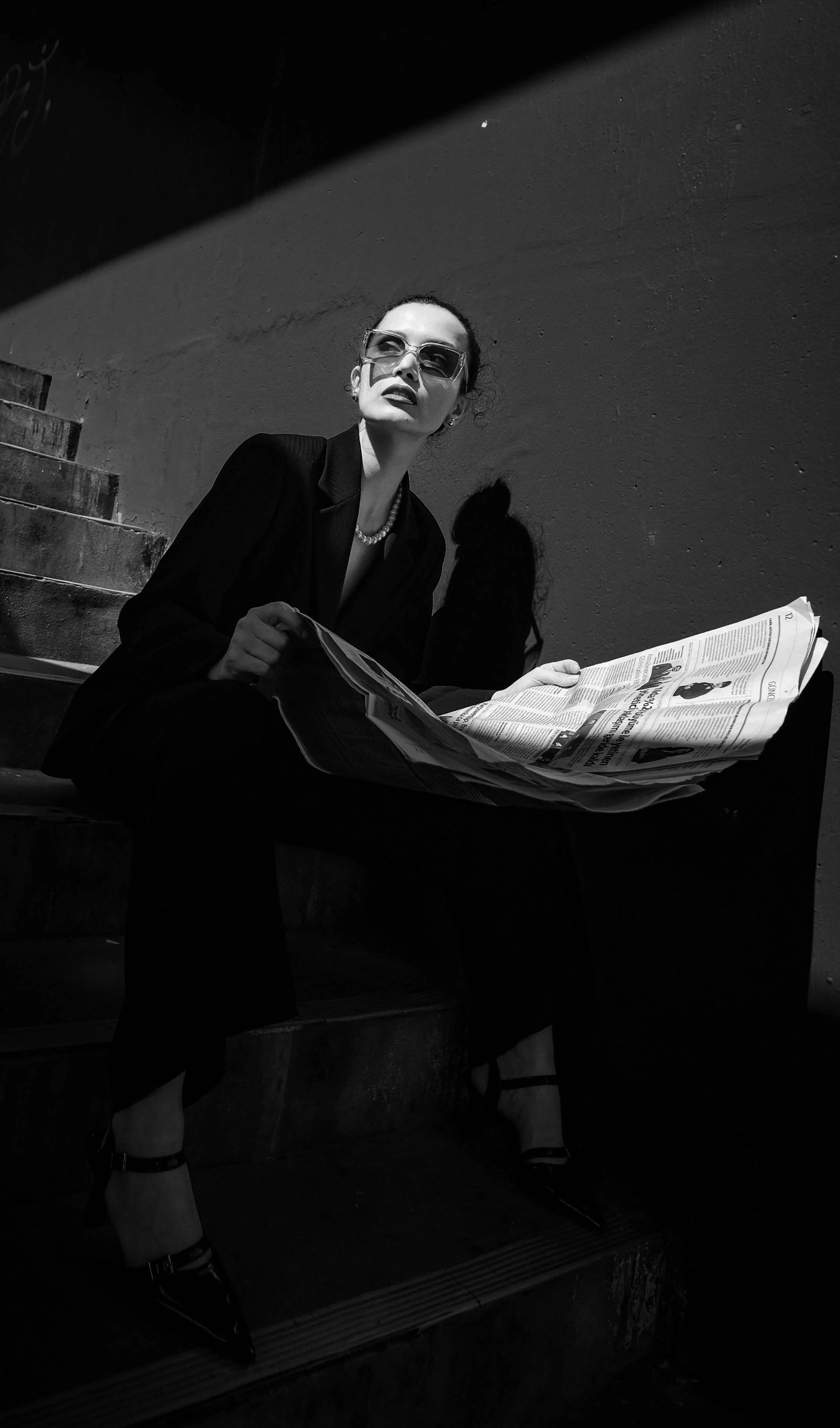 Black and white photo of a woman sitting on stairs, wearing sunglasses and a dark suit, reading a newspaper, with high heels and jewelry.