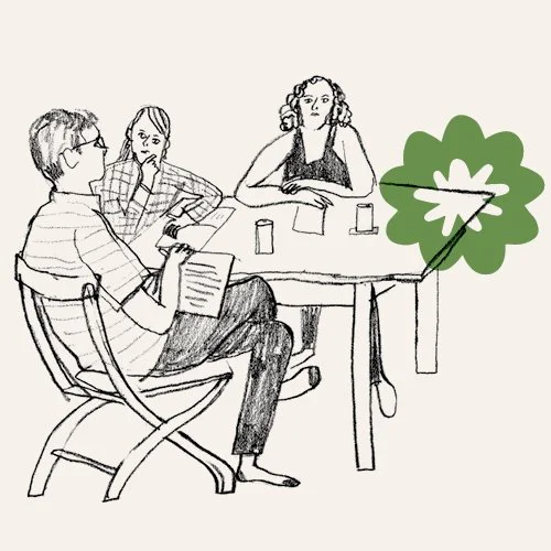 Three people sitting at a table, two are women and one person is man, in a discussion, with a large green leafy plant on the right.