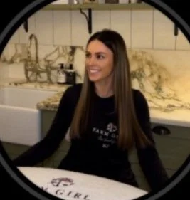 Woman with long brown hair smiling, sitting at a table in a restaurant or cafe, wearing a black shirt.