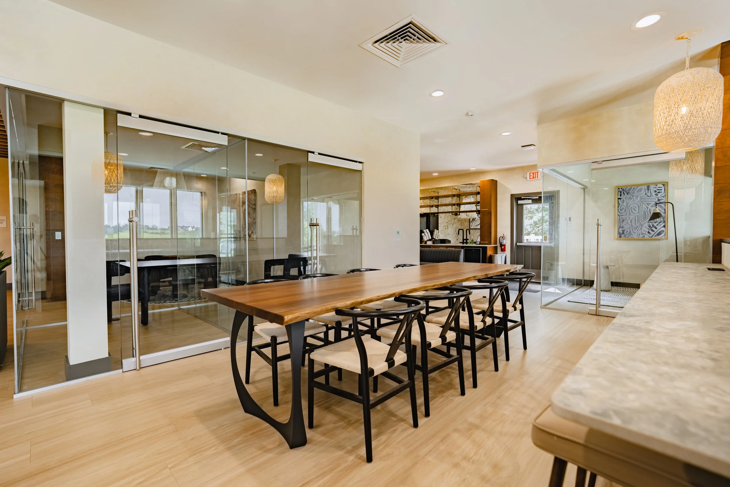 Lounge area with a long wooden table, black chairs, and glass-enclosed meeting rooms. Pendant lights hang from the ceiling, and there is a wall art piece on the right.