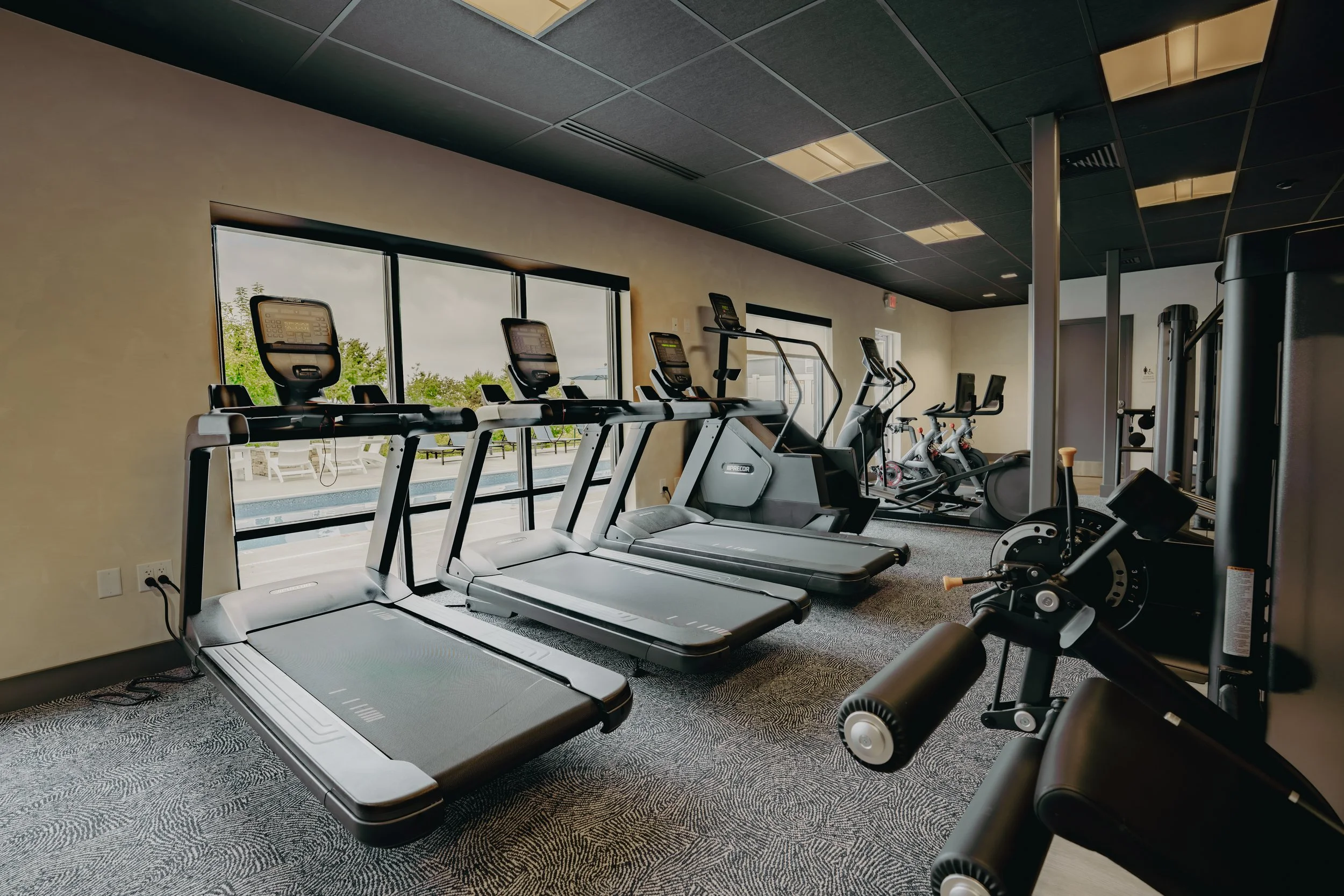 Gym room with treadmills and exercise bikes facing large windows, with outdoor pool and lounge chairs visible outside.
