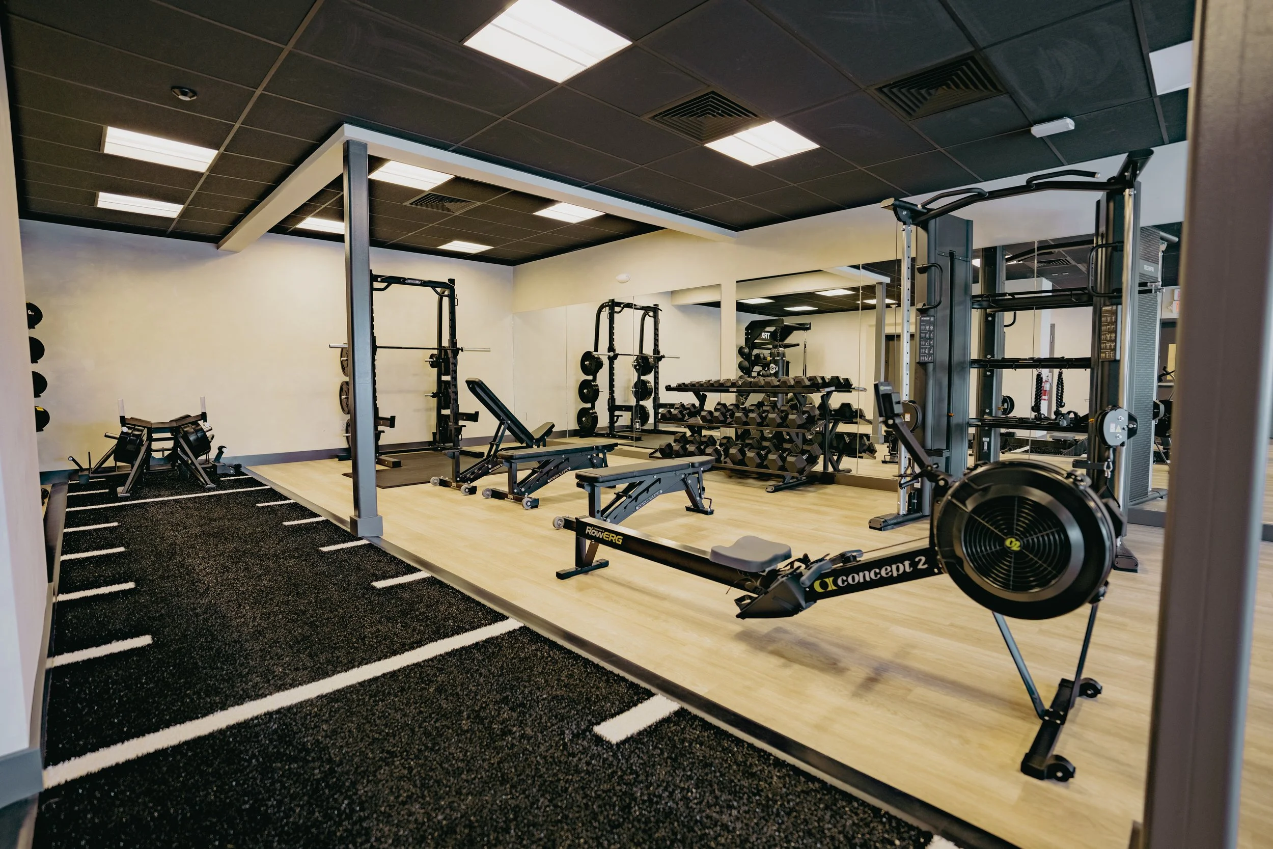 A gym with exercise equipment including rowing machines, weight racks, and benches, with a black and white striped rubber floor and a large mirror on the wall.