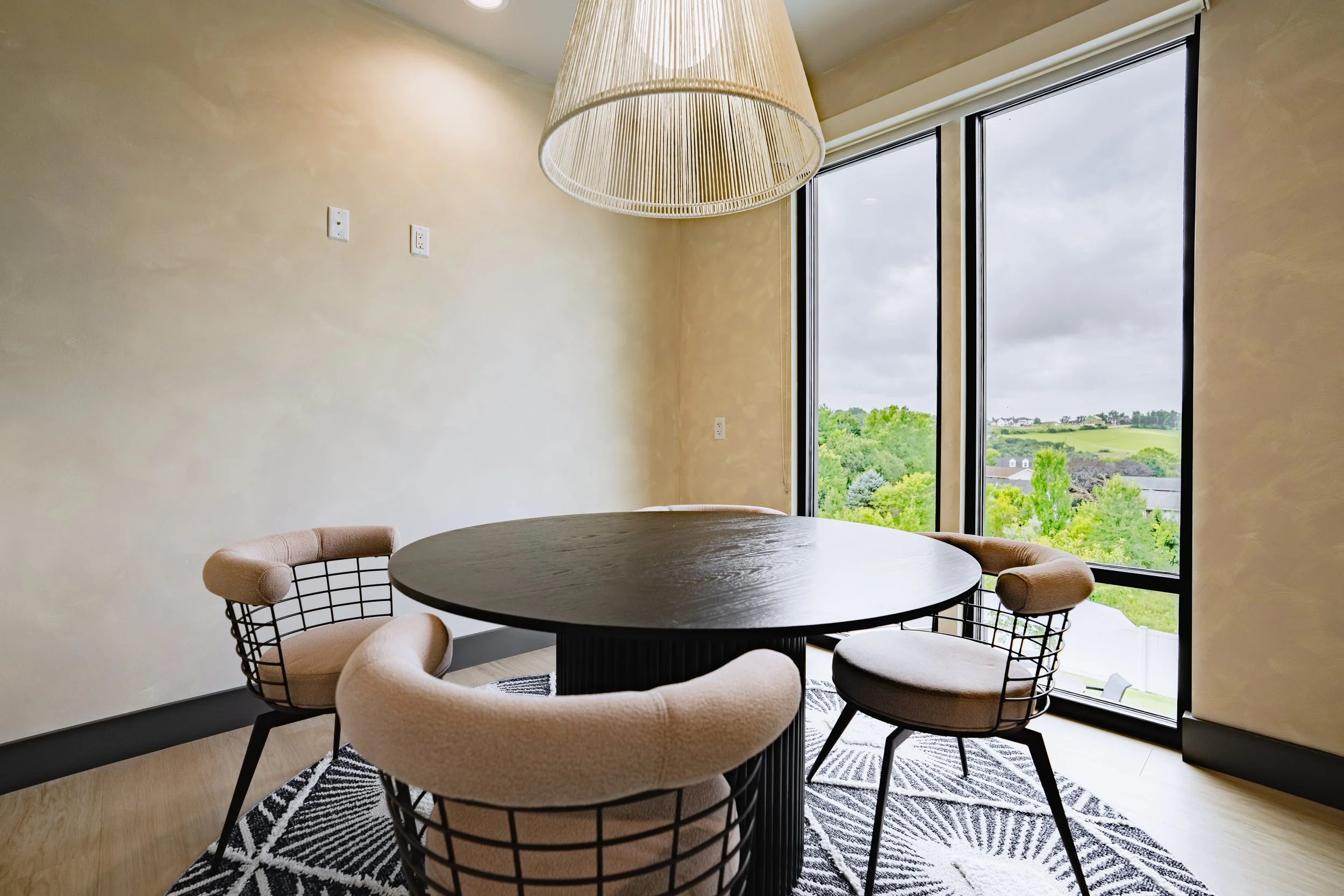 A round black table surrounded by four modern pink cushioned chairs with black wire frames, placed on a black and white patterned rug near large windows with a view of green trees and a cloudy sky.