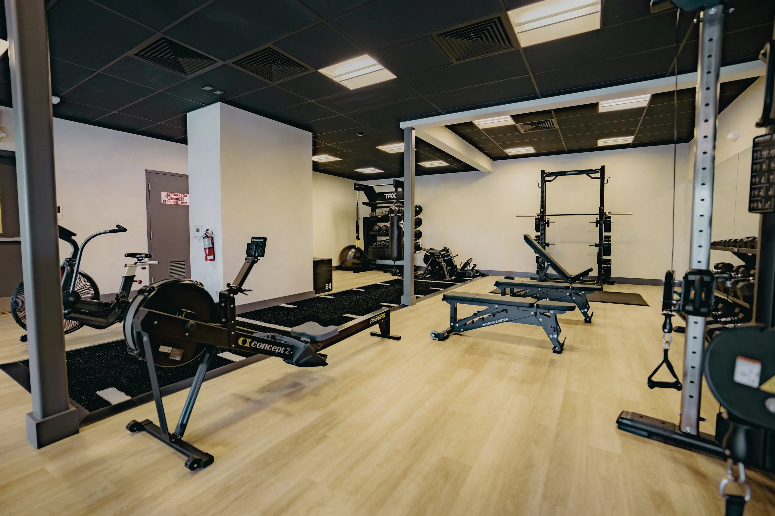 Empty gym with workout equipment including rowing machine, weight bench, squat rack, and various weights, with a black ceiling and wooden floor.