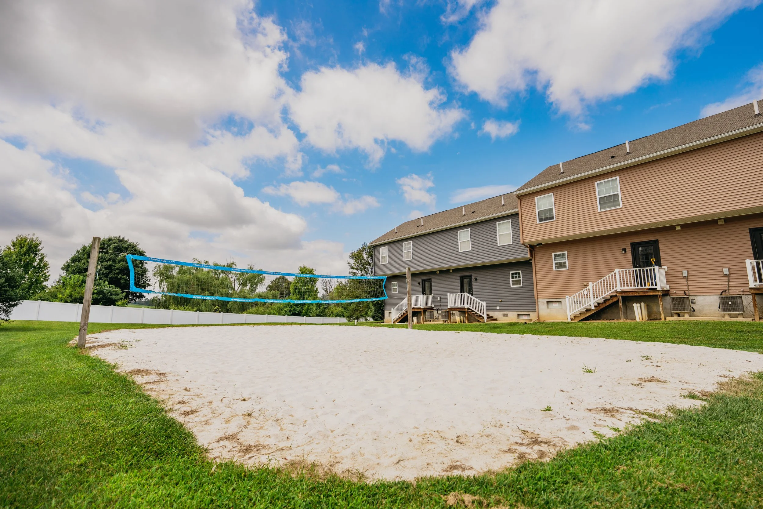 A backyard with a volleyball net set up on a sandy court, bordered by green grass. In the background, there are brown and gray residential buildings with stairs leading to doors, and a partly cloudy sky overhead.