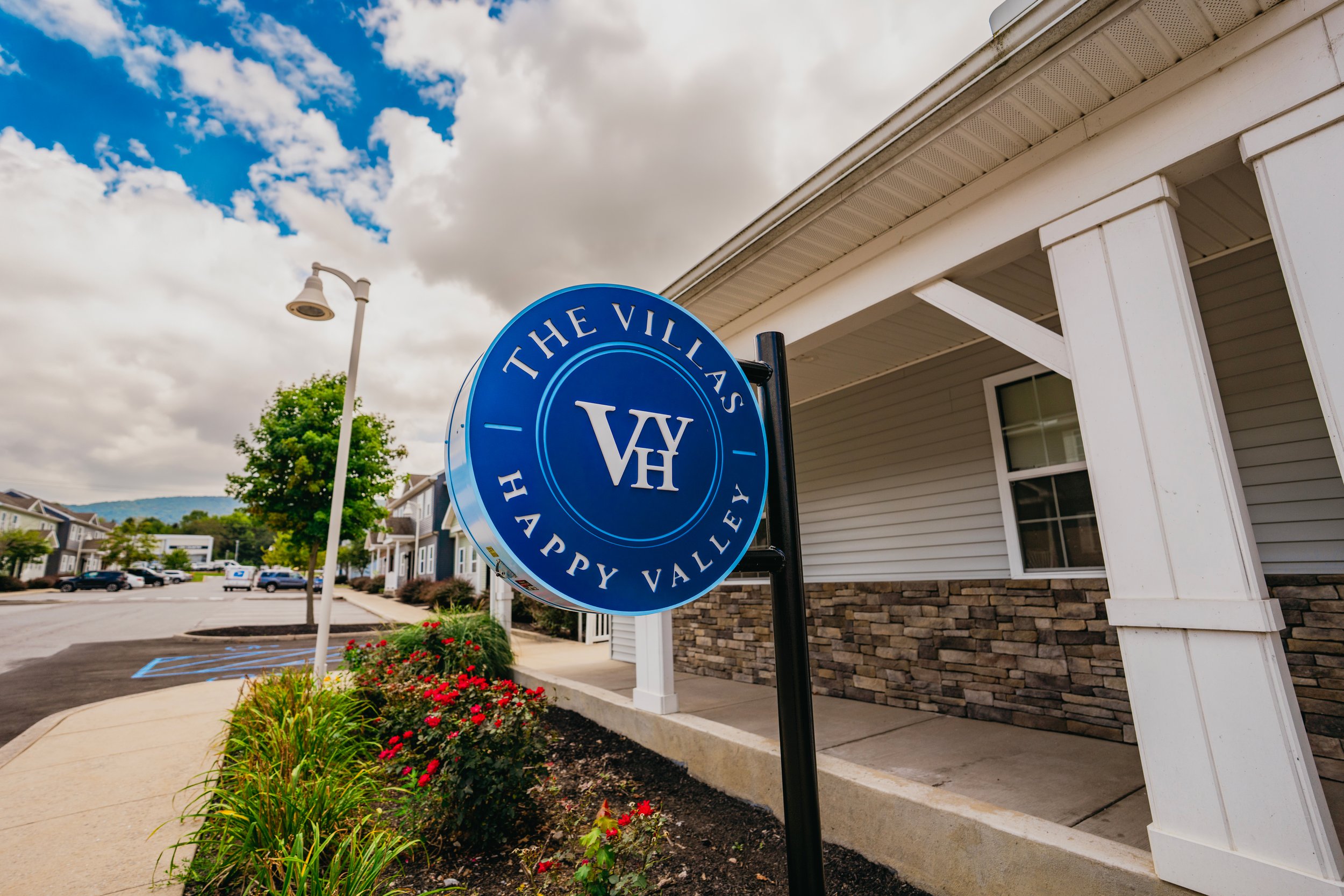 Sign for The Villages Happy Valley neighborhood in a suburban area with residential buildings, a streetlight, some parking spaces, and landscaping with flowers and bushes under partly cloudy skies.