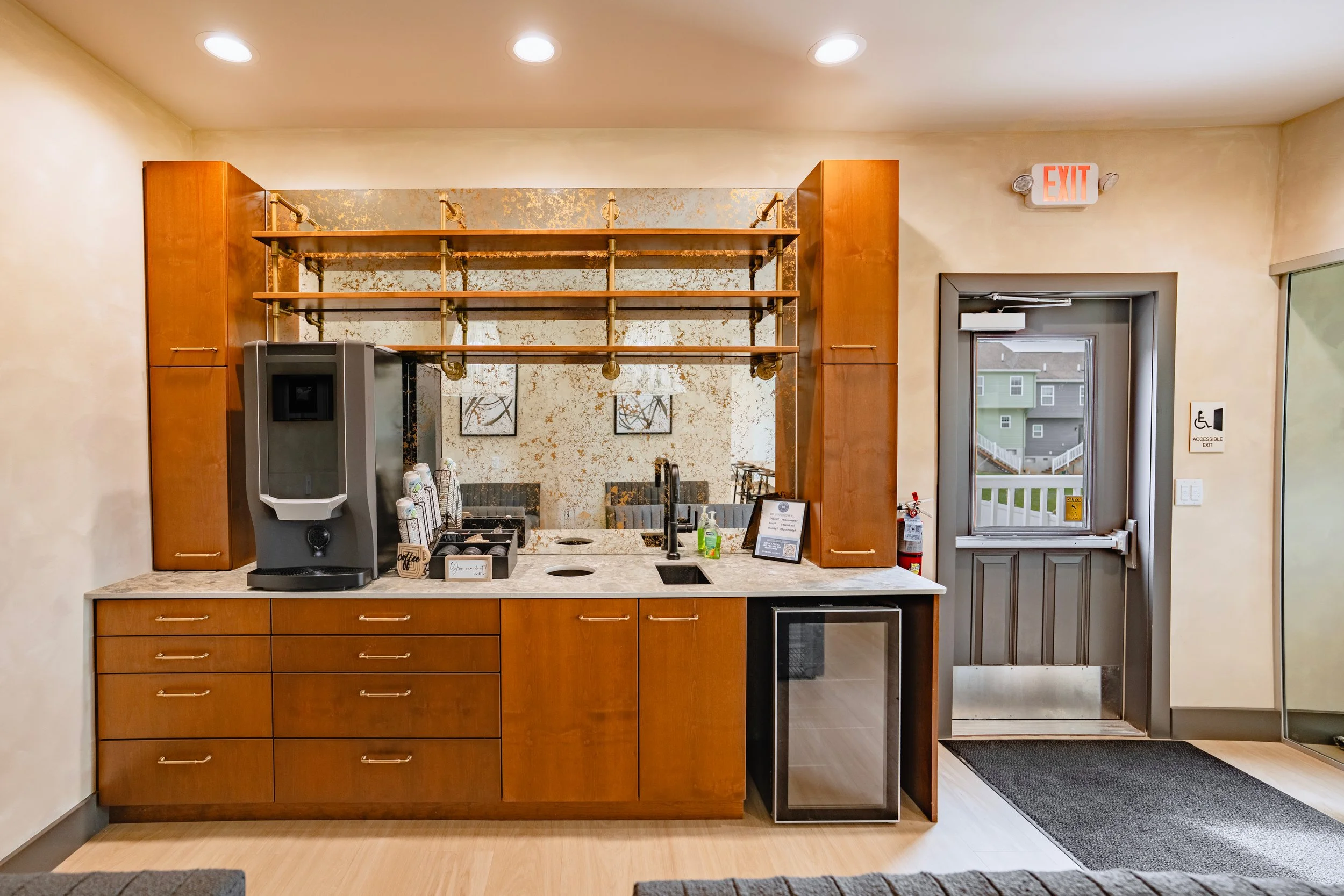 Indoor coffee  counter with wooden cabinets, granite countertop, coffee machine, and hand sanitizer bottles. A wall with a gold-and-silver decorative pattern and framed artwork.