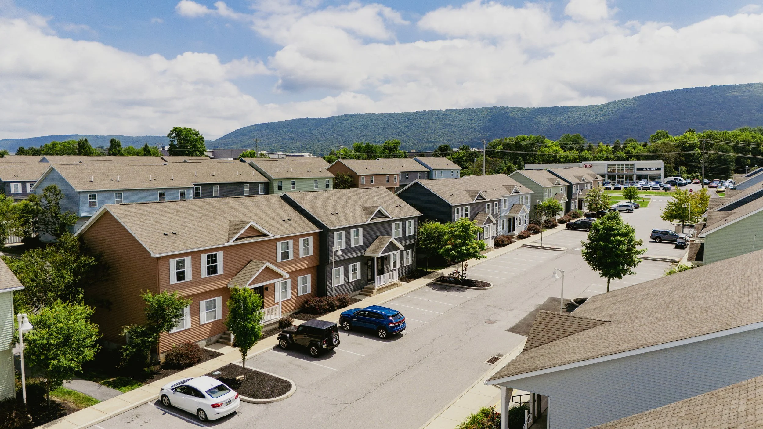 View of a residential neighborhood with colorful houses, a parking lot, and green trees, with forested mountains in the background under a partly cloudy sky.