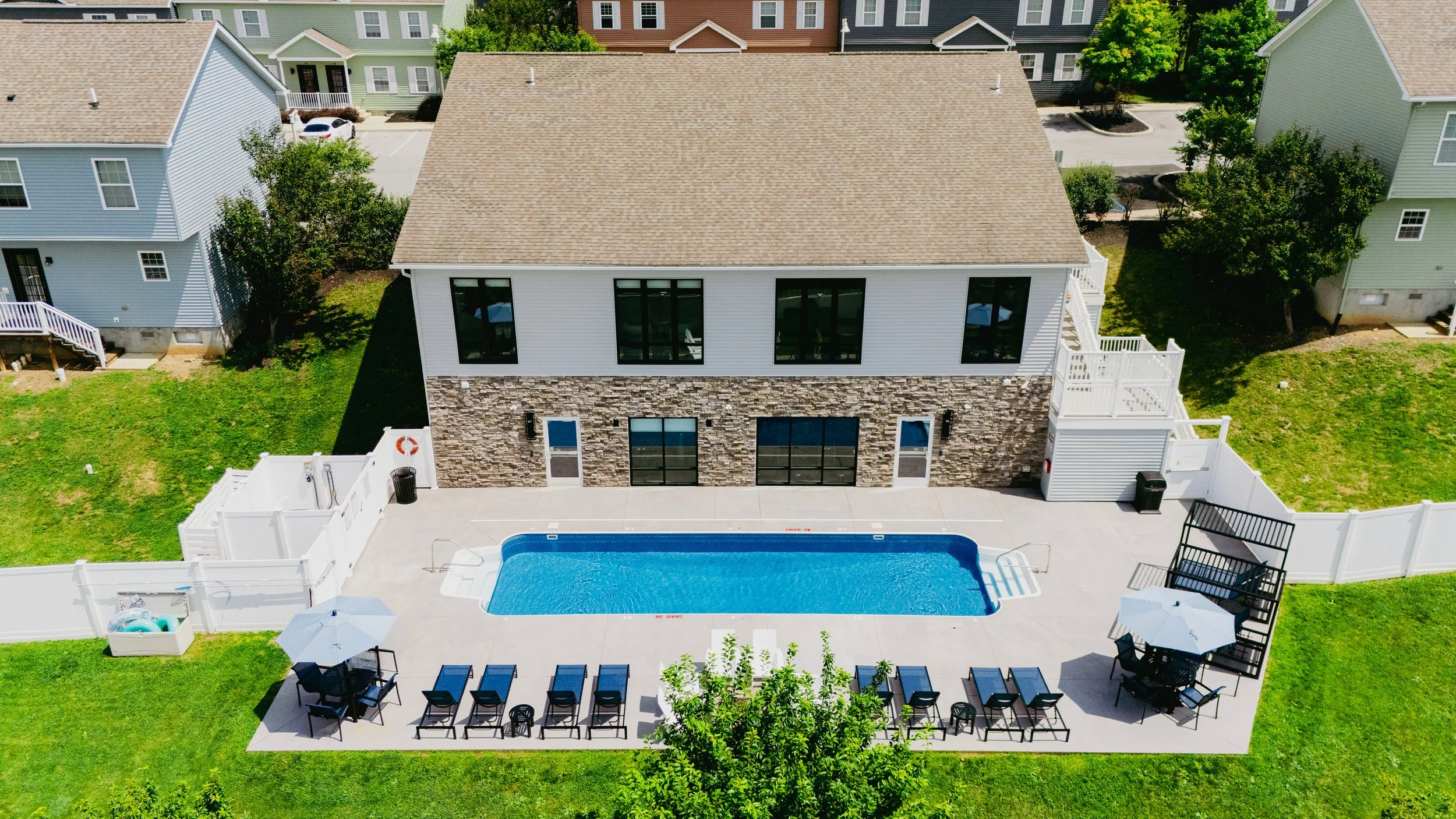 Aerial view of a modern house with a stone and white siding exterior, a beige gabled roof, large black windows, a backyard with a swimming pool, patio area with chairs and umbrella, surrounded by green grass and neighboring houses.