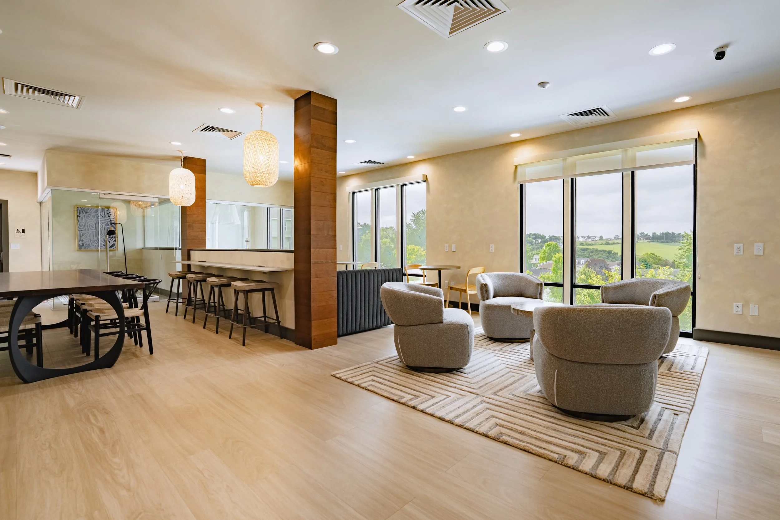 Bright and modern lounge area with large windows overlooking greenery, beige walls, and light wood flooring. Cherry wood accents, hanging pendant lights, and a mix of armchairs and tables create a comfortable atmosphere.