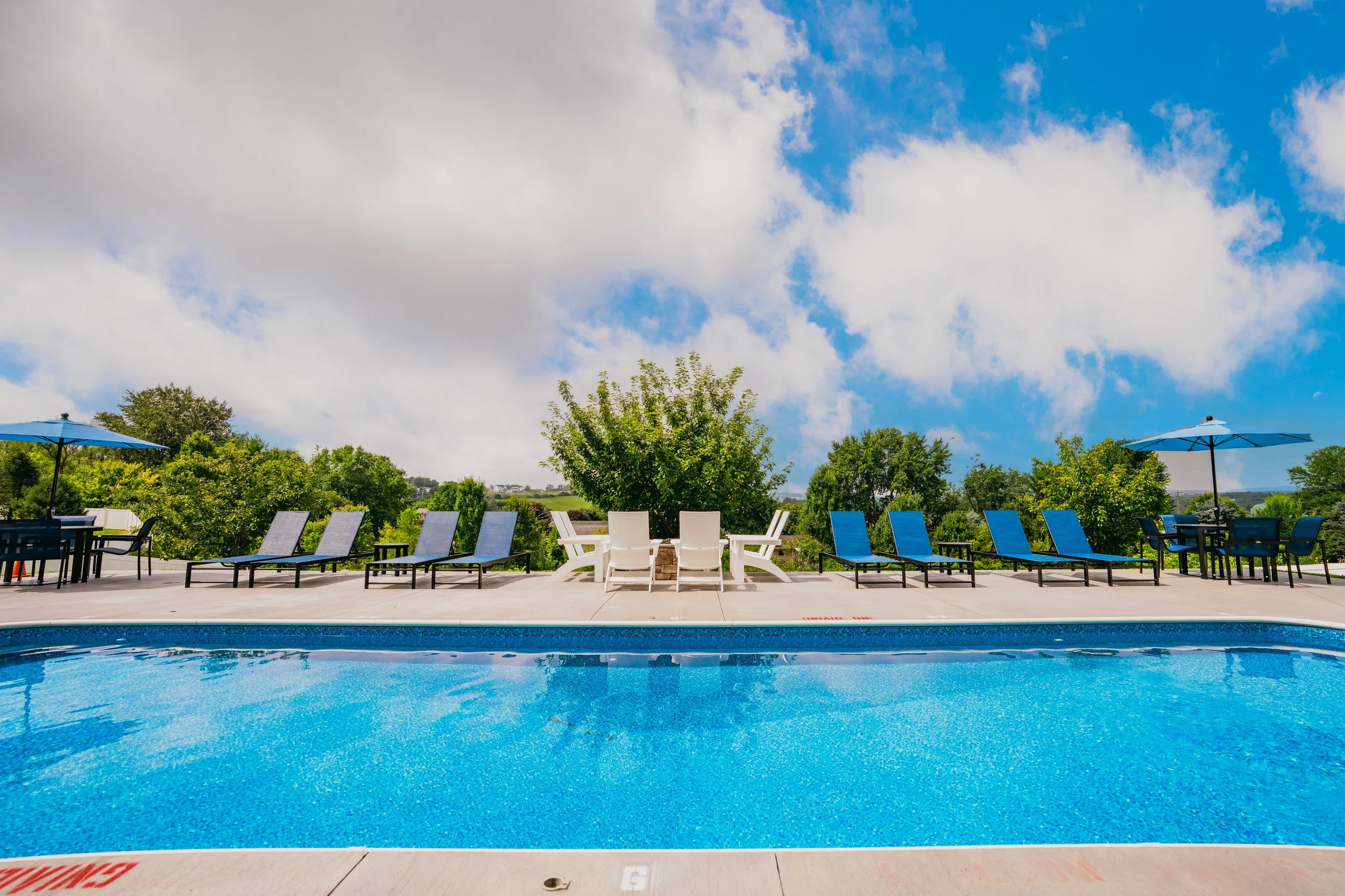 An outdoor swimming pool area with lounge chairs and umbrellas, surrounded by trees and greenery, under a partly cloudy blue sky.