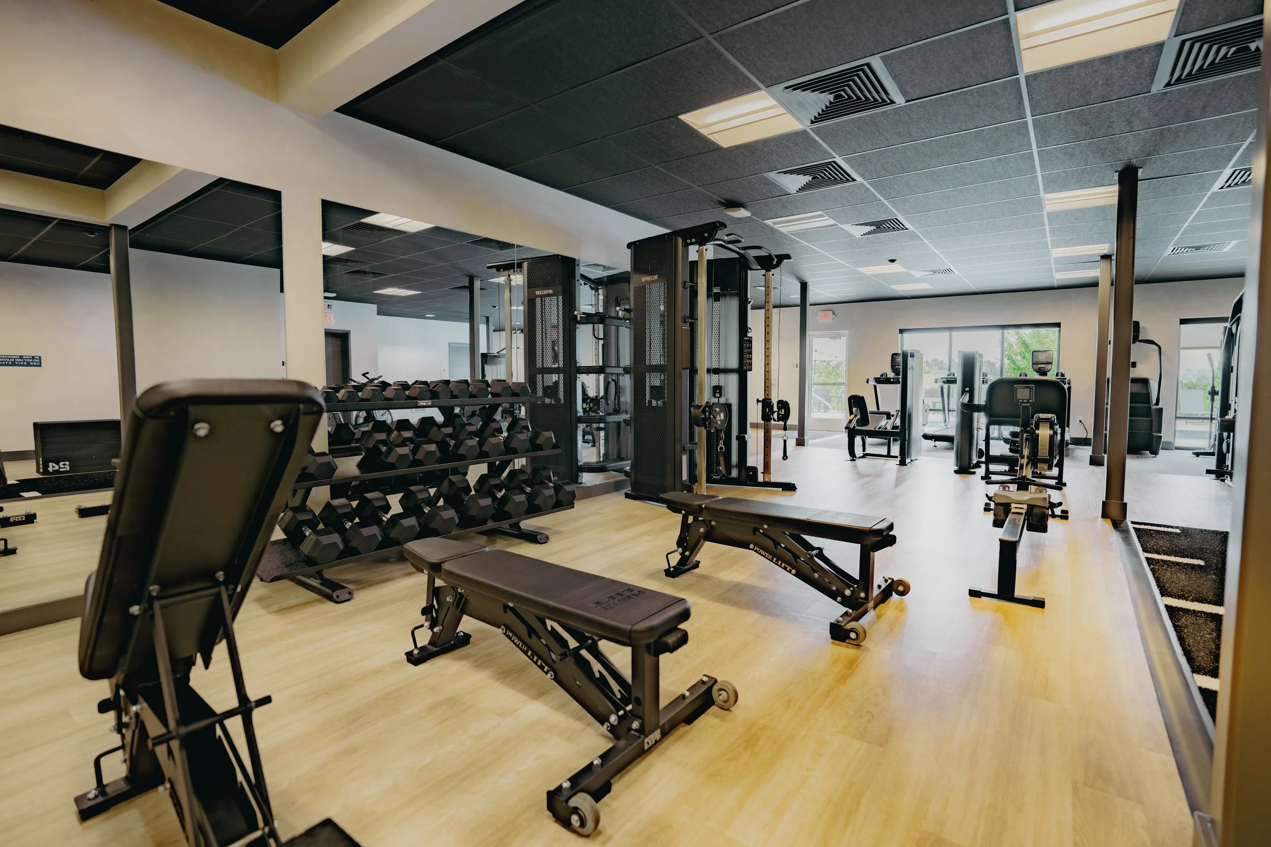 A gym with various workout equipment including dumbbells, weightlifting machines, benches, and cardio machines, illuminated by ceiling lights with large windows in the background.