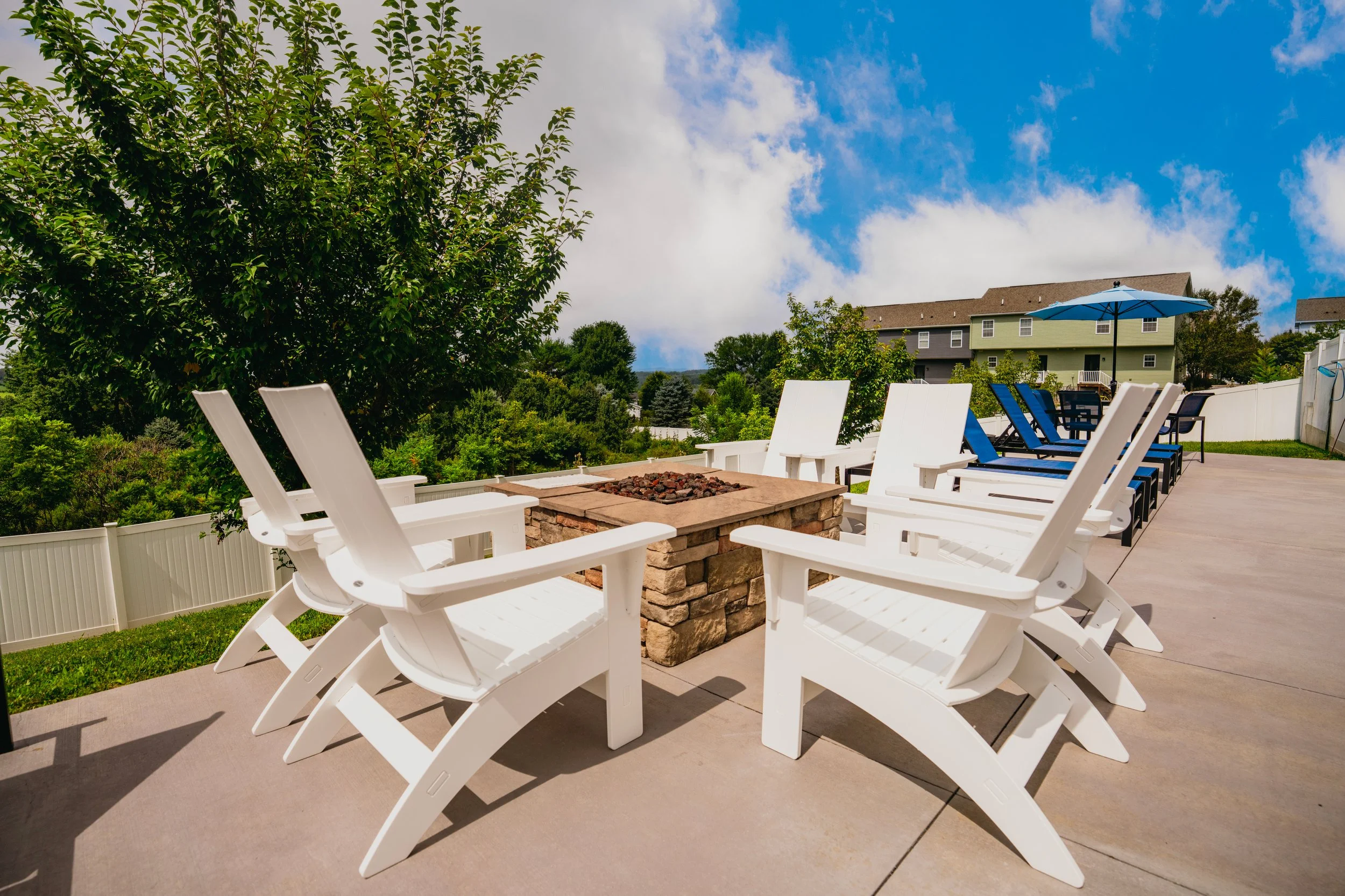 A backyard patio with white Adirondack chairs, a stone fire pit, and an umbrella. There are trees, bushes, and a house with green siding and multiple windows in the background, under a partly cloudy sky.
