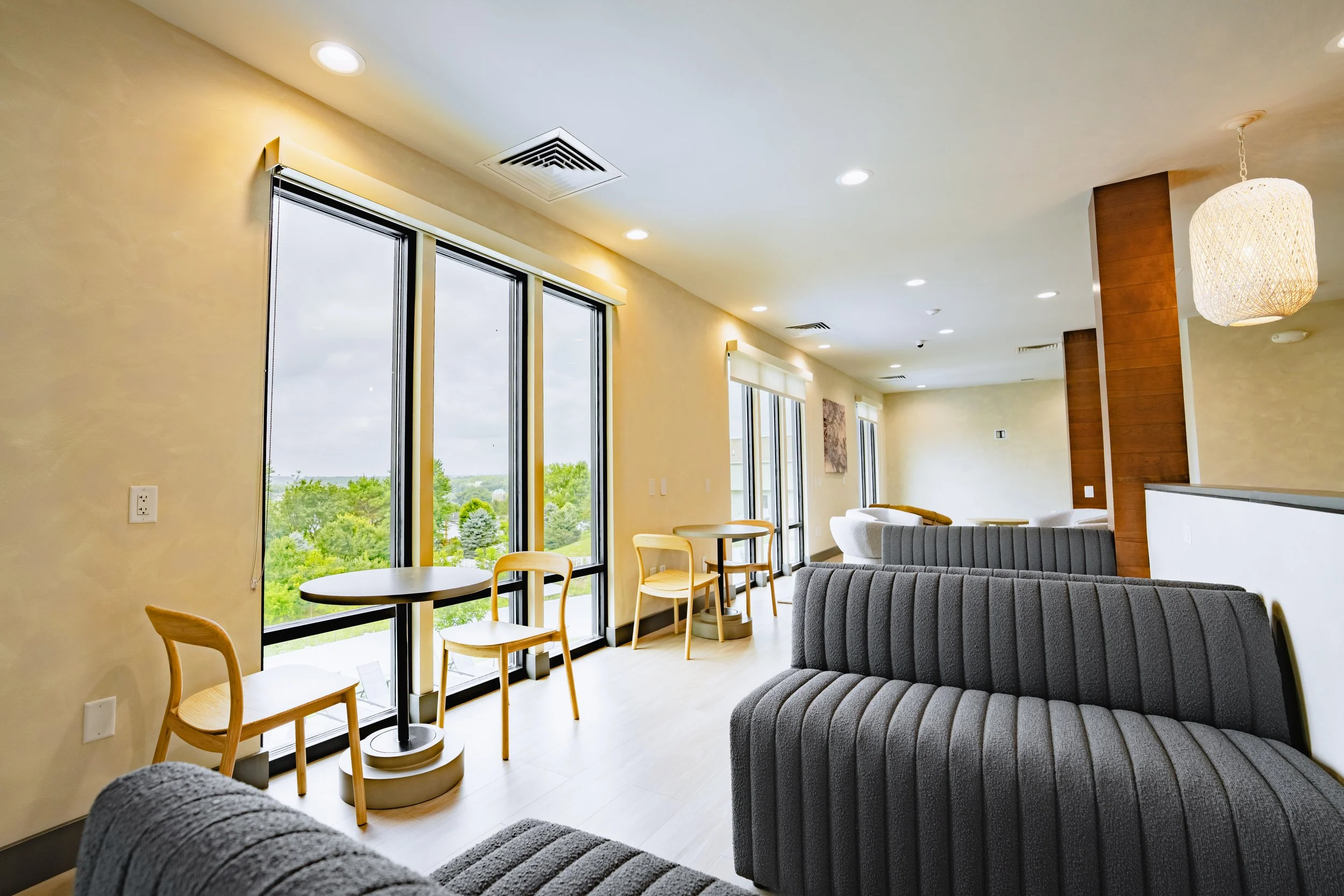 Interior of a modern lounge area with floor-to-ceiling windows, wooden chairs, a small round table, and a gray upholstered sofa. There is a hanging light fixture and trees visible outside the windows.
