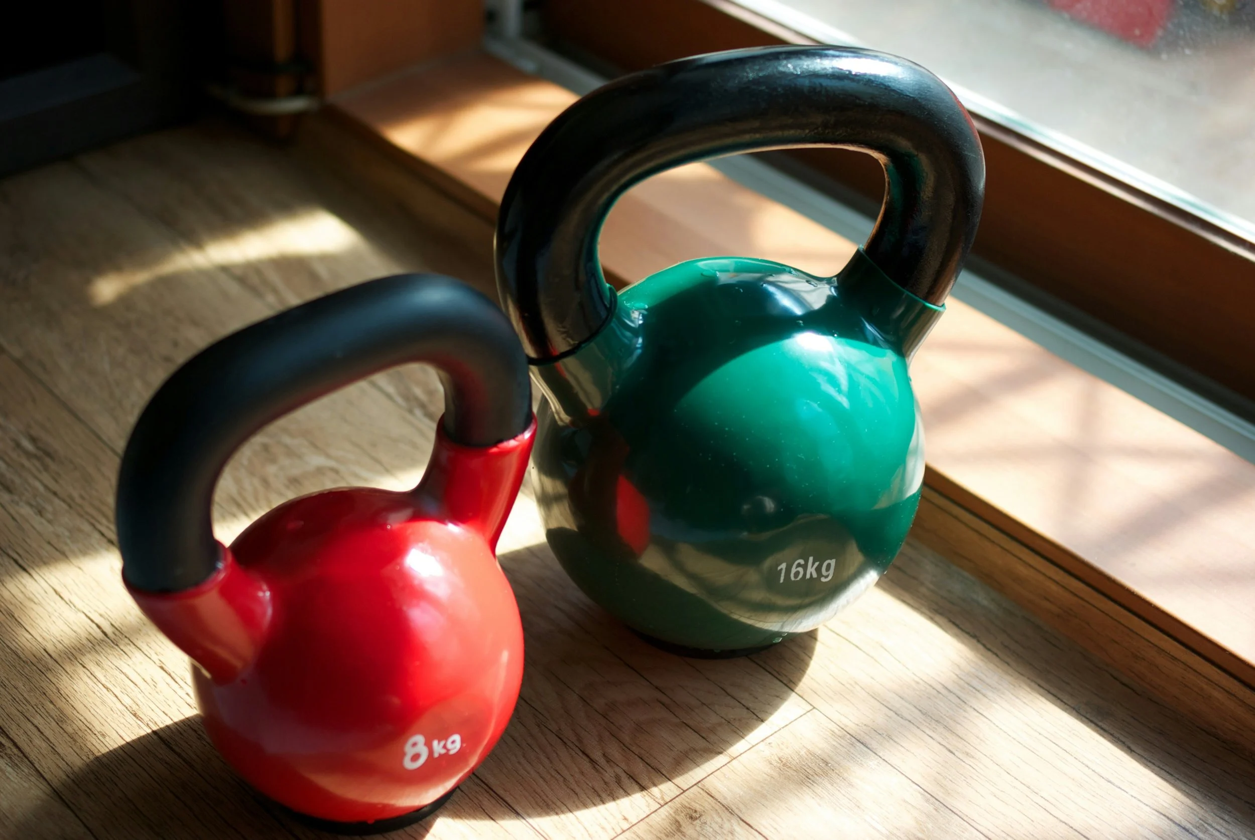 Two kettlebells, one red labeled 8kg and one green labeled 16kg, placed on a wooden floor near a window with sunlight and shadows.