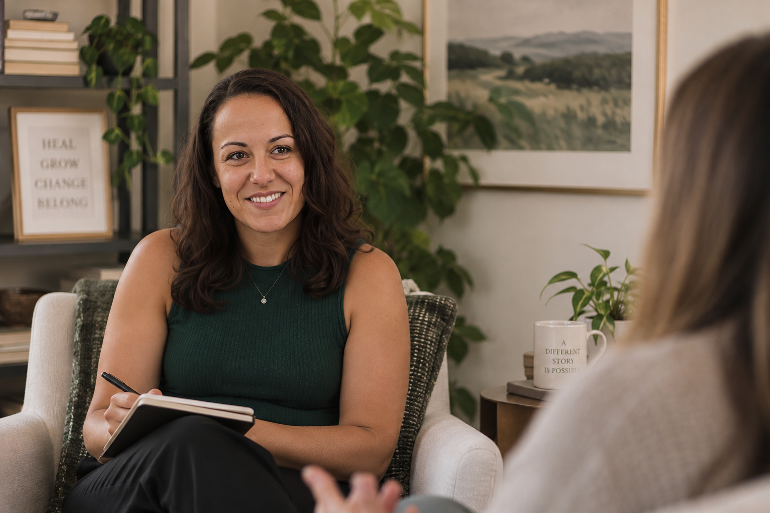 A woman in a green sleeveless top smiling while taking notes during a counseling session in a cozy room with plants and motivational signs.