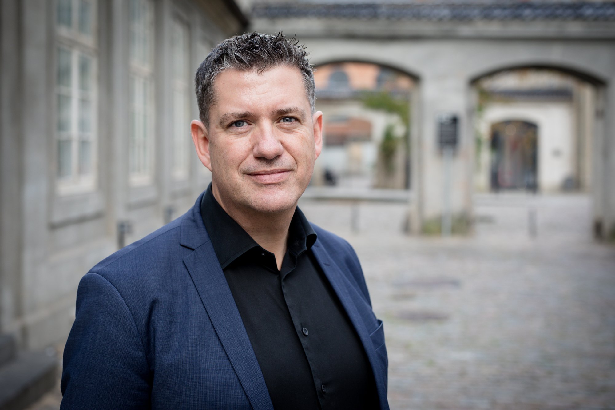 A man in a dark blue suit and black shirt standing outdoors with blurred buildings in the background.