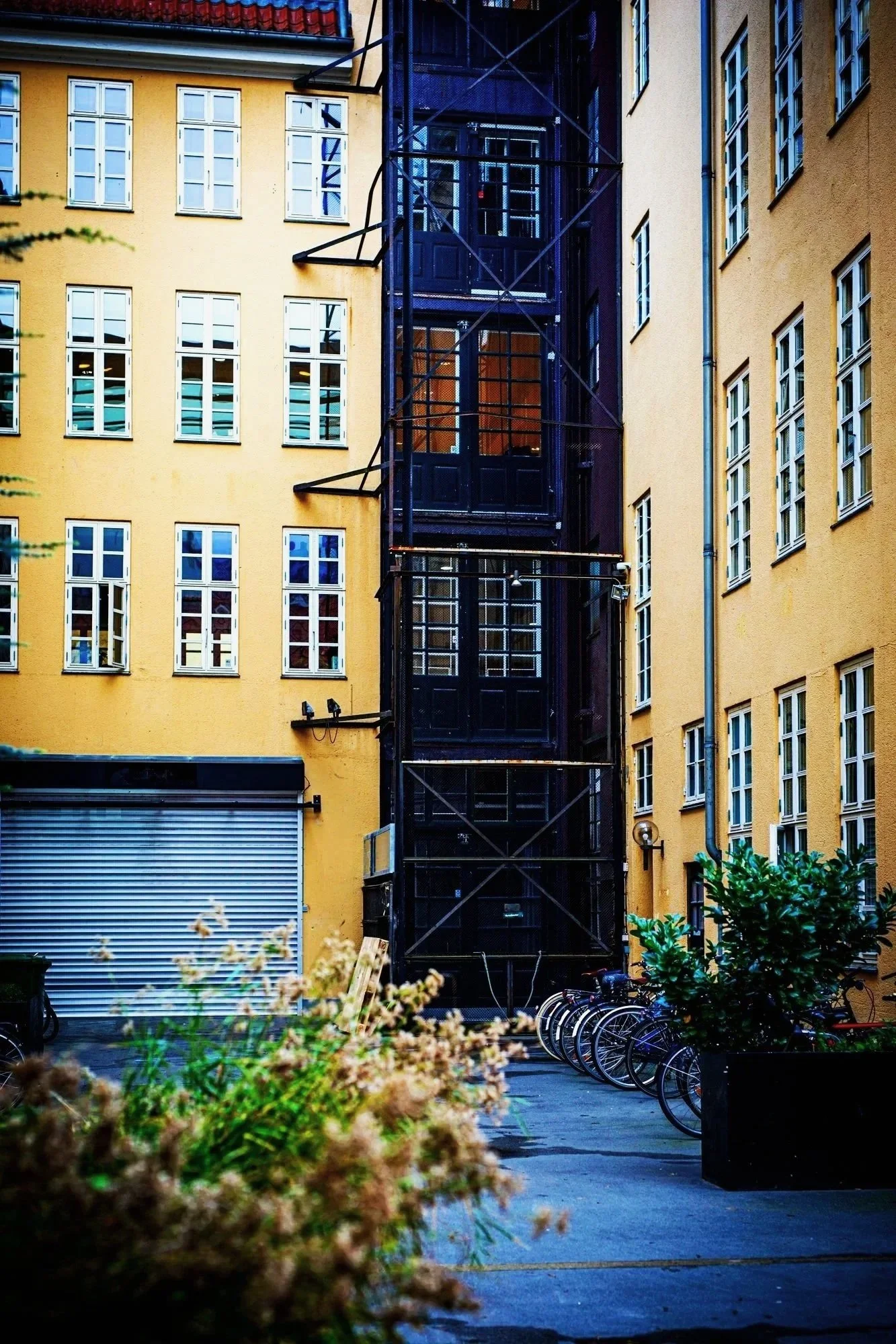 An outdoor urban scene with yellow apartment buildings, a black external elevator, bicycles, and potted plants.