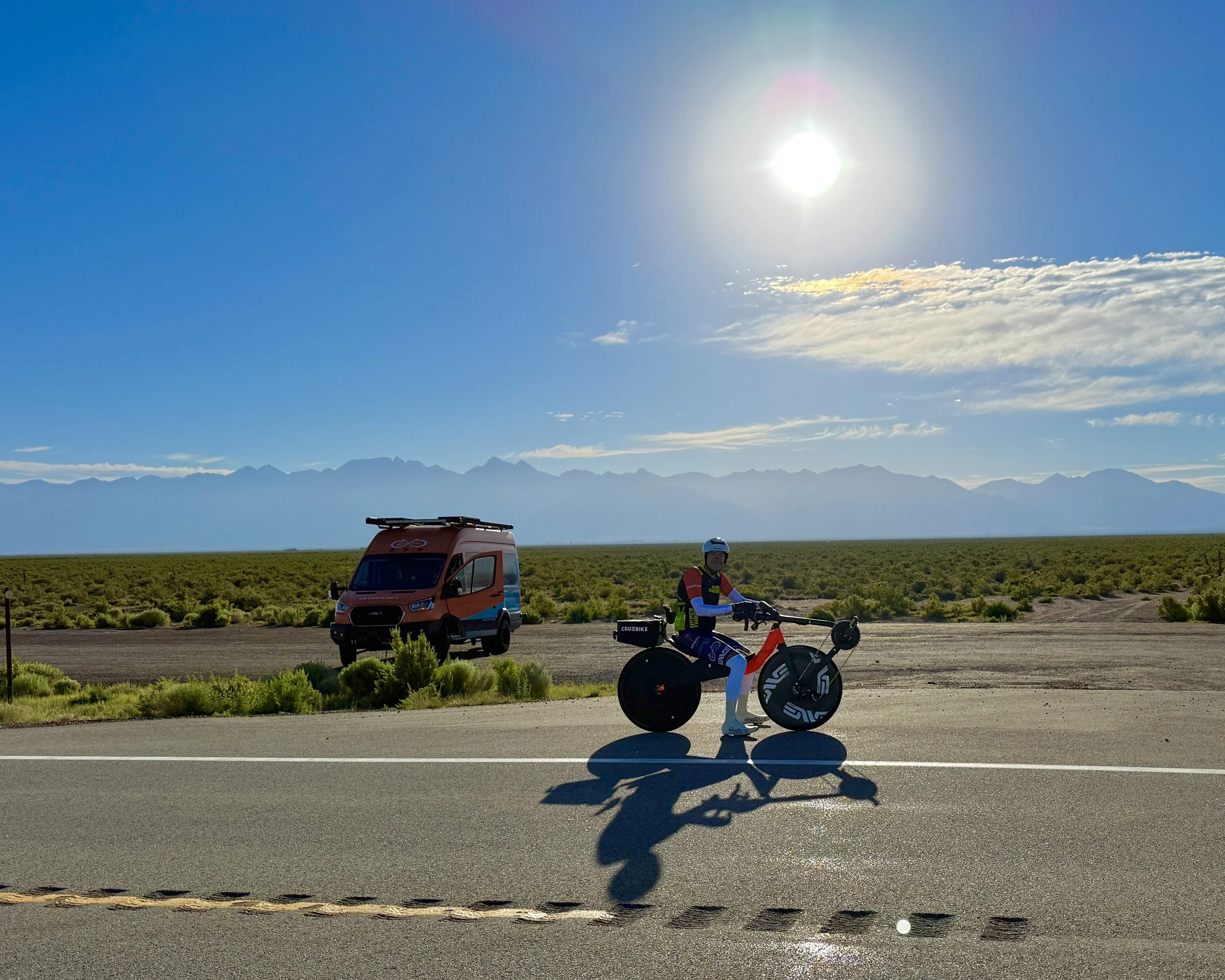 Matthew Lefthand sitting up on his recumbent bicycle posed on the side of the road in front of an orange and blue van with mountains in the background.