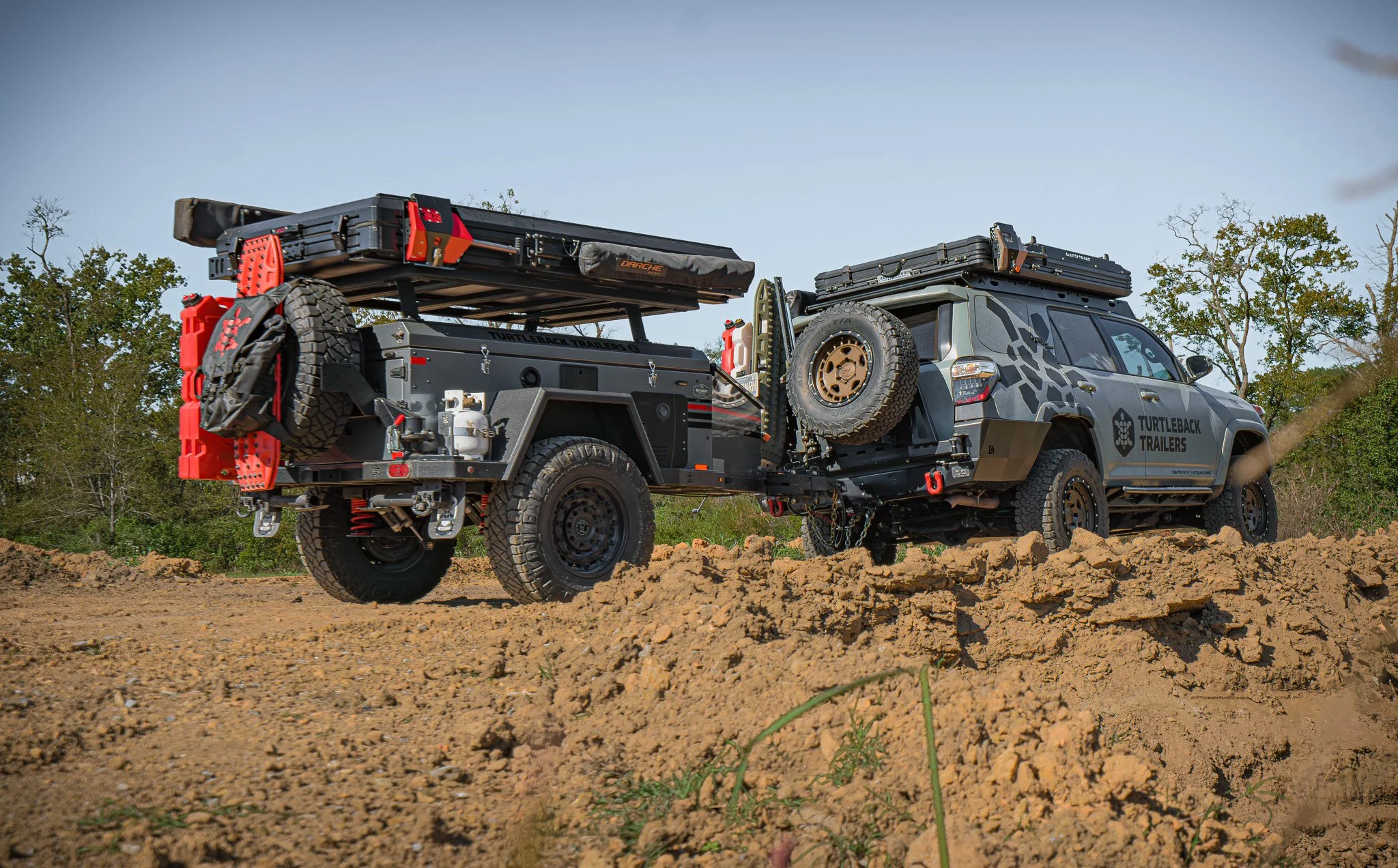 Off-road vehicle with trailer on uneven dirt terrain, outdoors with trees in the background.
