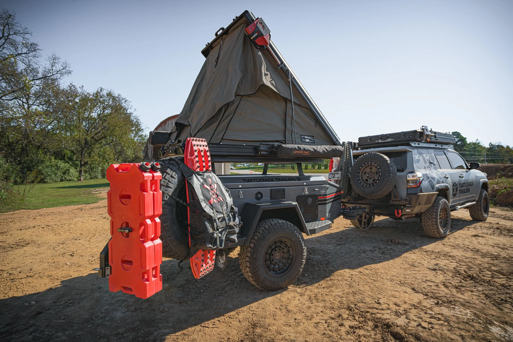 Off-road trailer attached to a pickup truck with a rooftop tent, spare tire, and storage gear in an outdoor setting.