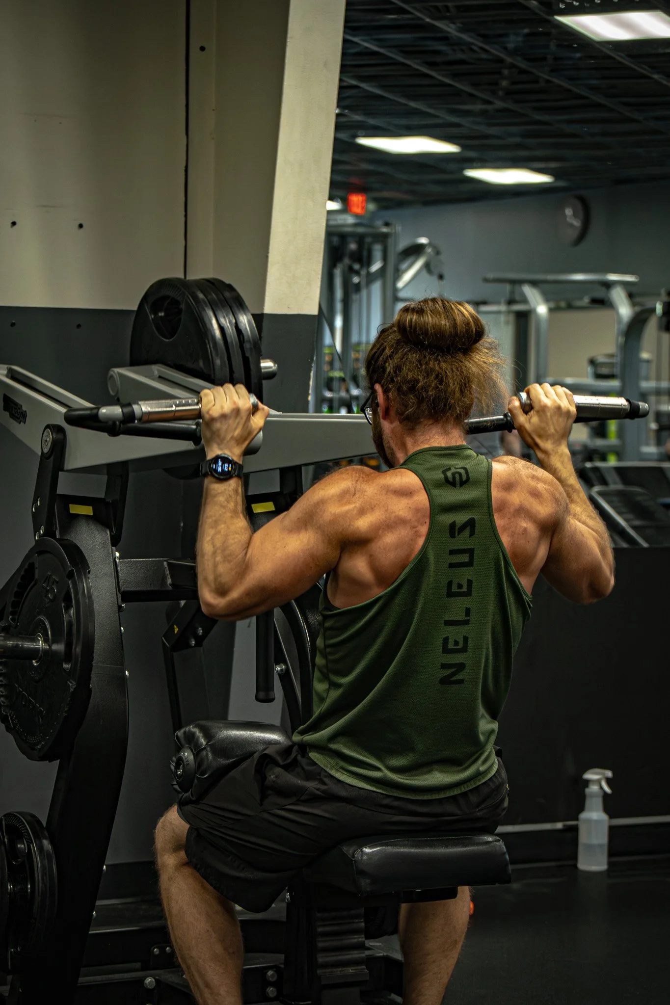 A man with muscular arms and brown hair tied in a bun is doing a shoulder press exercise in a gym, sitting on a bench and lifting a barbell with weights.