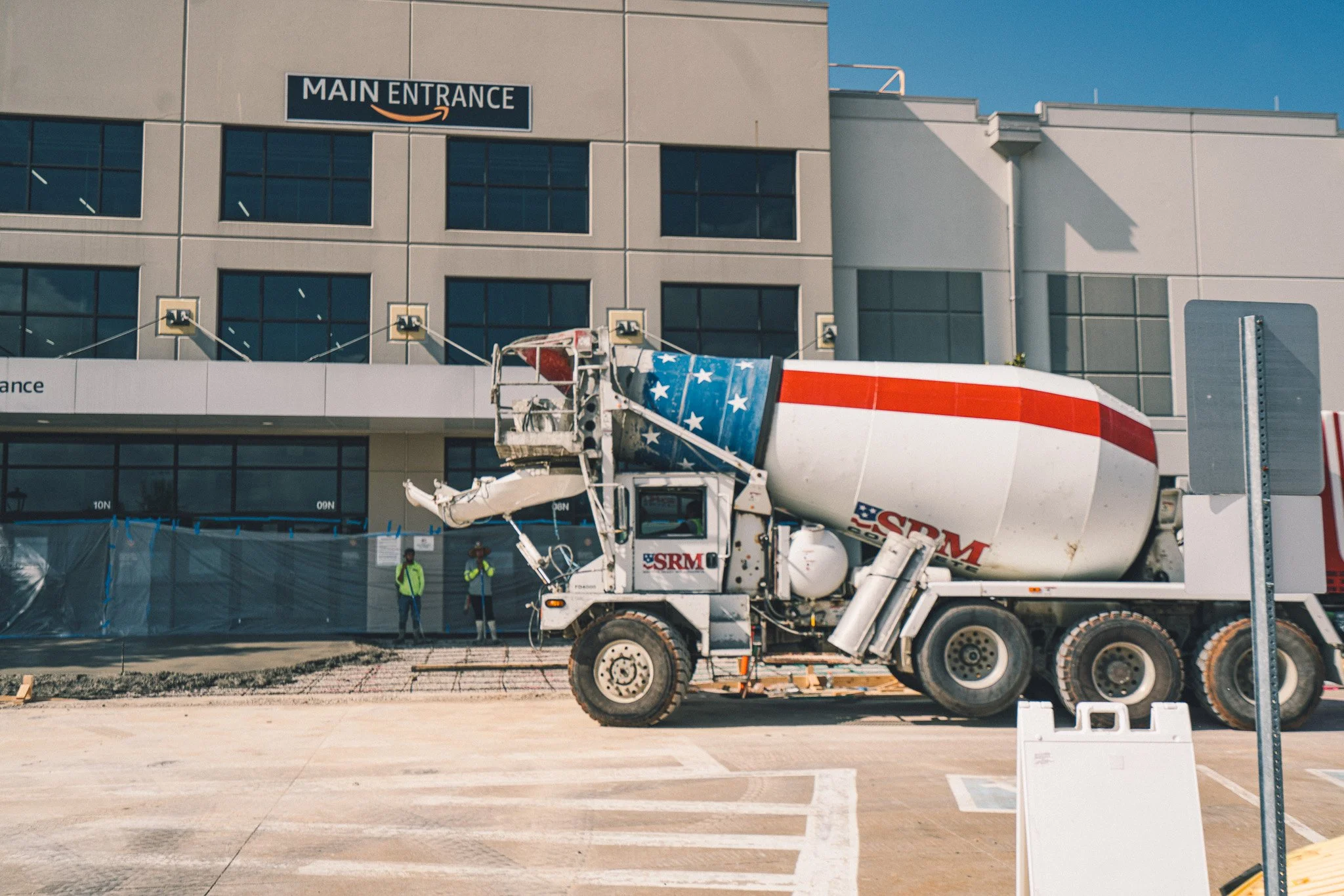 Concrete mixer truck with an American flag design on the drum, working outside a building with a sign reading 'Main Entrance.'