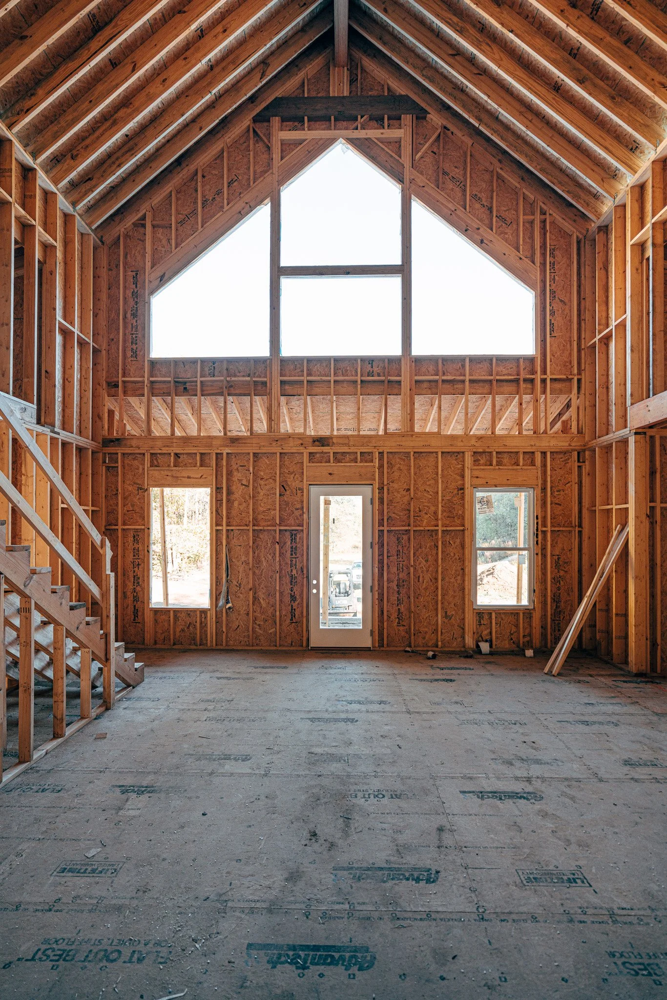 Interior of a house under construction, showing exposed wooden framing, a large triangular window at the top, two smaller windows on either side of a door at the bottom, and unfinished flooring.
