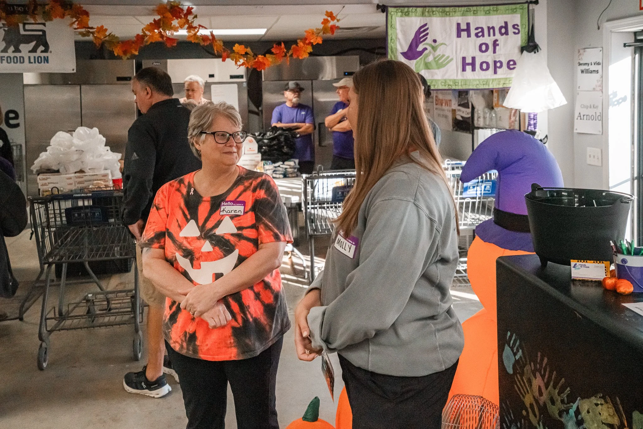 Two women, one wearing a tie-dye Halloween shirt with a pumpkin face, and the other in a gray sweatshirt, are having a conversation in a community kitchen or event space. Both wear name tags, and there are decorations including orange autumn leaves and a poster that says 'Hands of Hope' in the background.