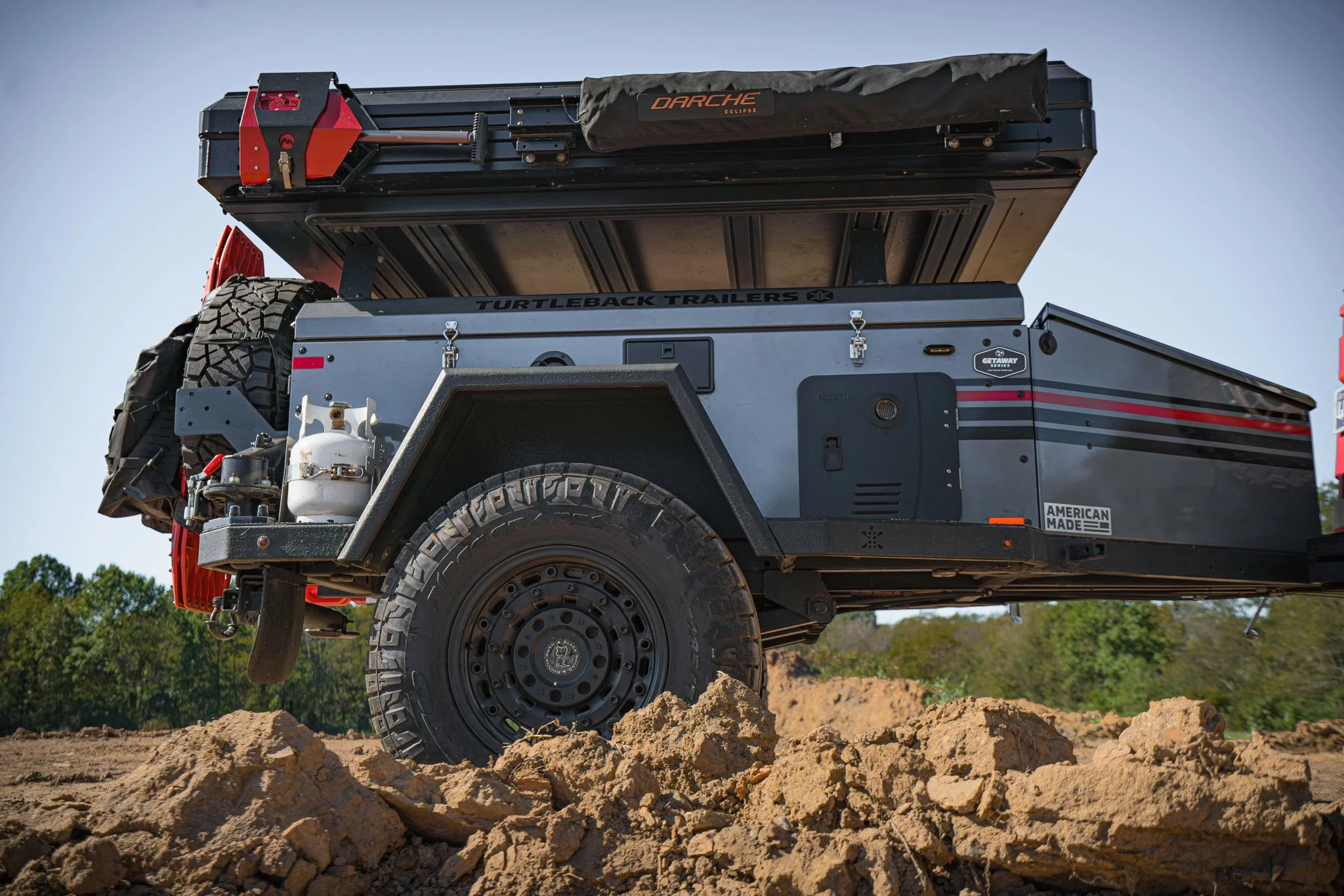 Off-road trailer with a mounted off-road vehicle on rough terrain, carrying a spare tire, propane tank, and equipment, under a clear sky.