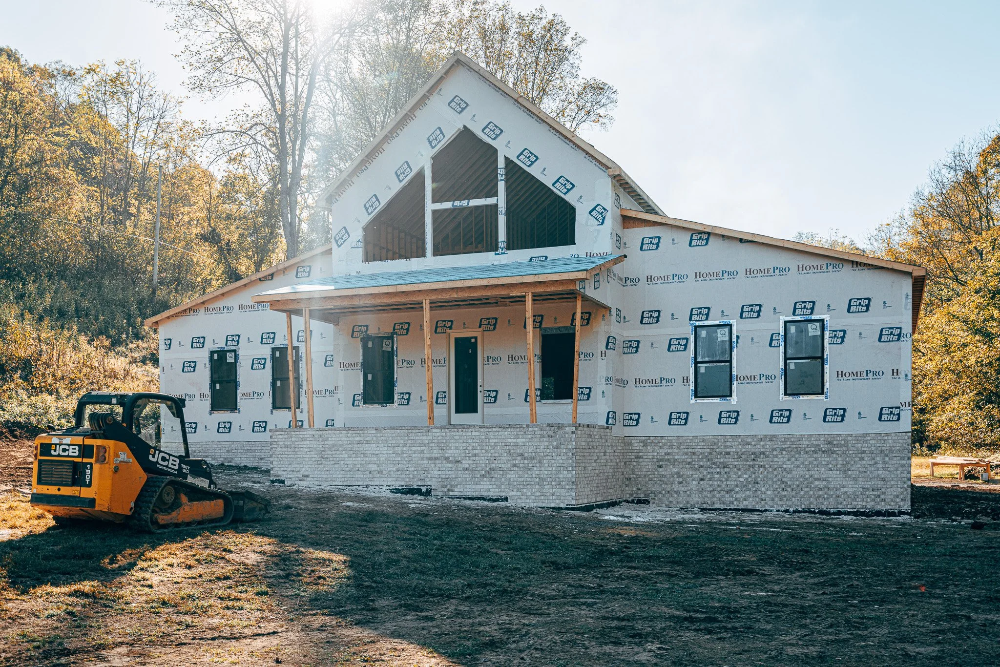 Under construction house with insulation and windows installed on a foundation, surrounded by trees and a small construction vehicle.