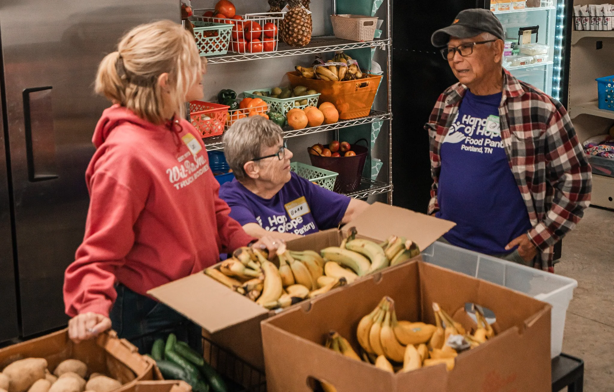 Three people at a food pantry stand around a table with bananas and other produce, engaged in conversation, with shelving of fruits and vegetables behind them.