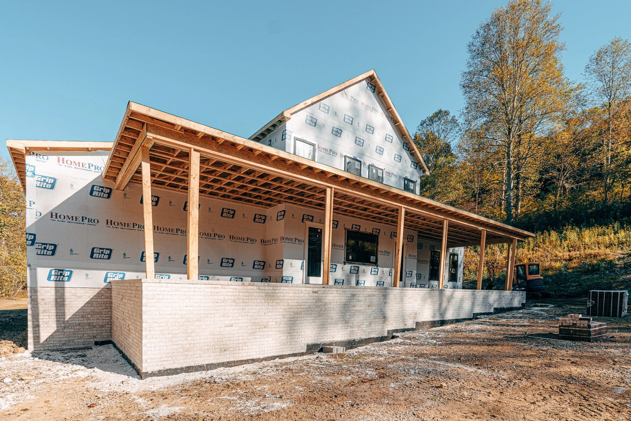 Under construction house with wooden framework, brick foundation, and protective house wrap, situated outdoors with trees in the background.