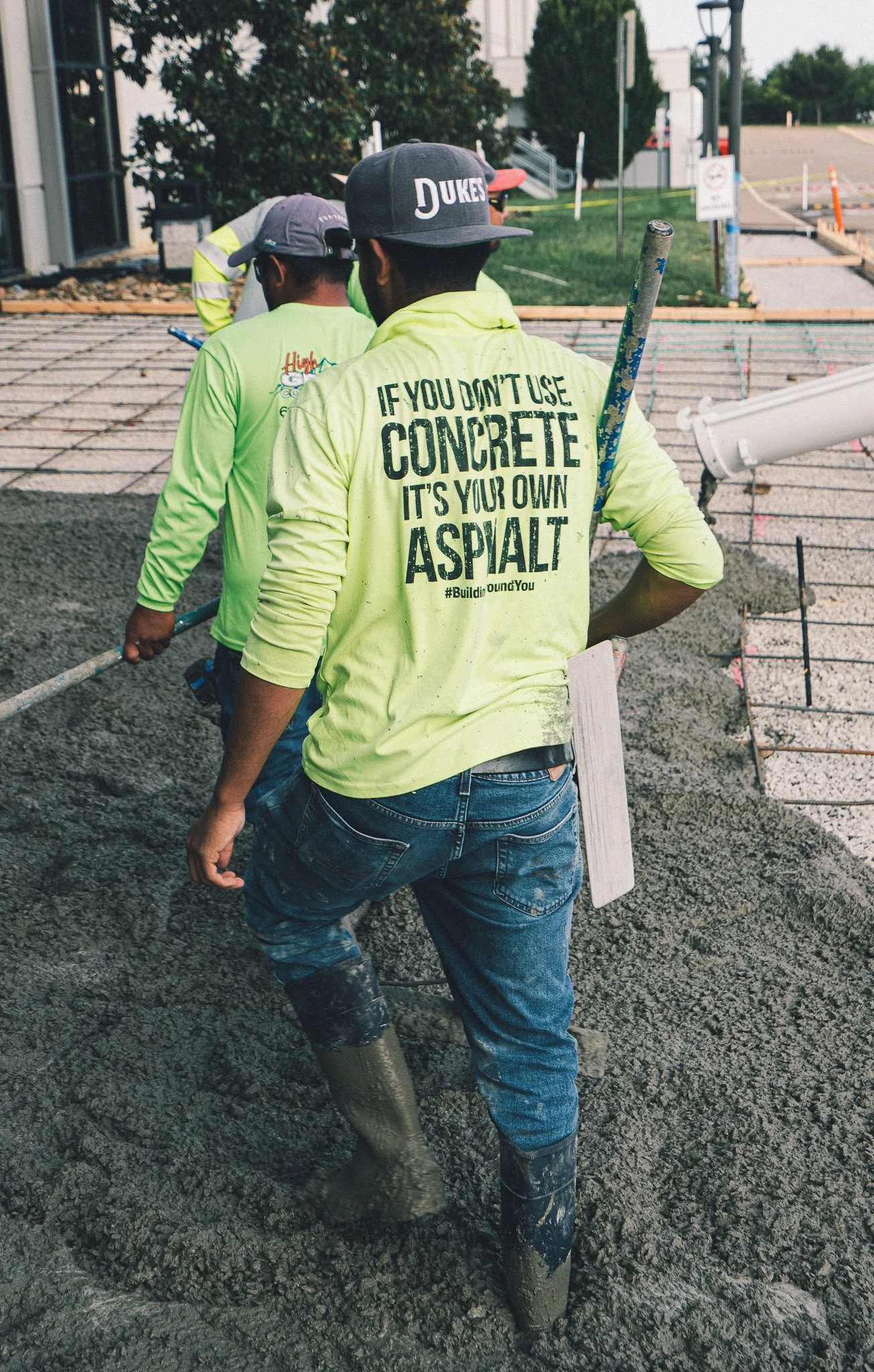 Construction workers pouring and leveling concrete on a building site, with one worker wearing a yellow hoodie that reads, 'If you don't use concrete, it's your own asphalt.'