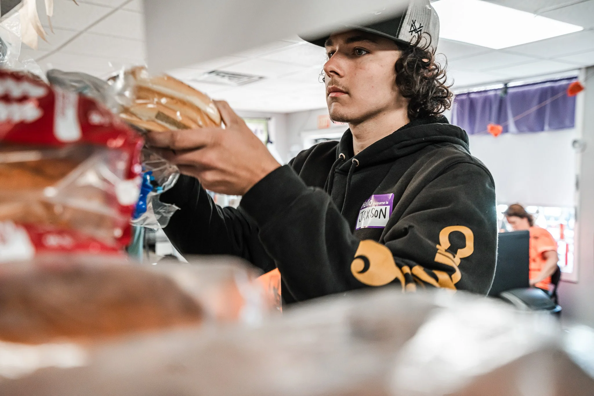 A young man with curly hair, wearing a black hoodie with orange text and a gray cap, organizes bread on a shelf inside a store. He has a name tag that reads 'Jackson.'